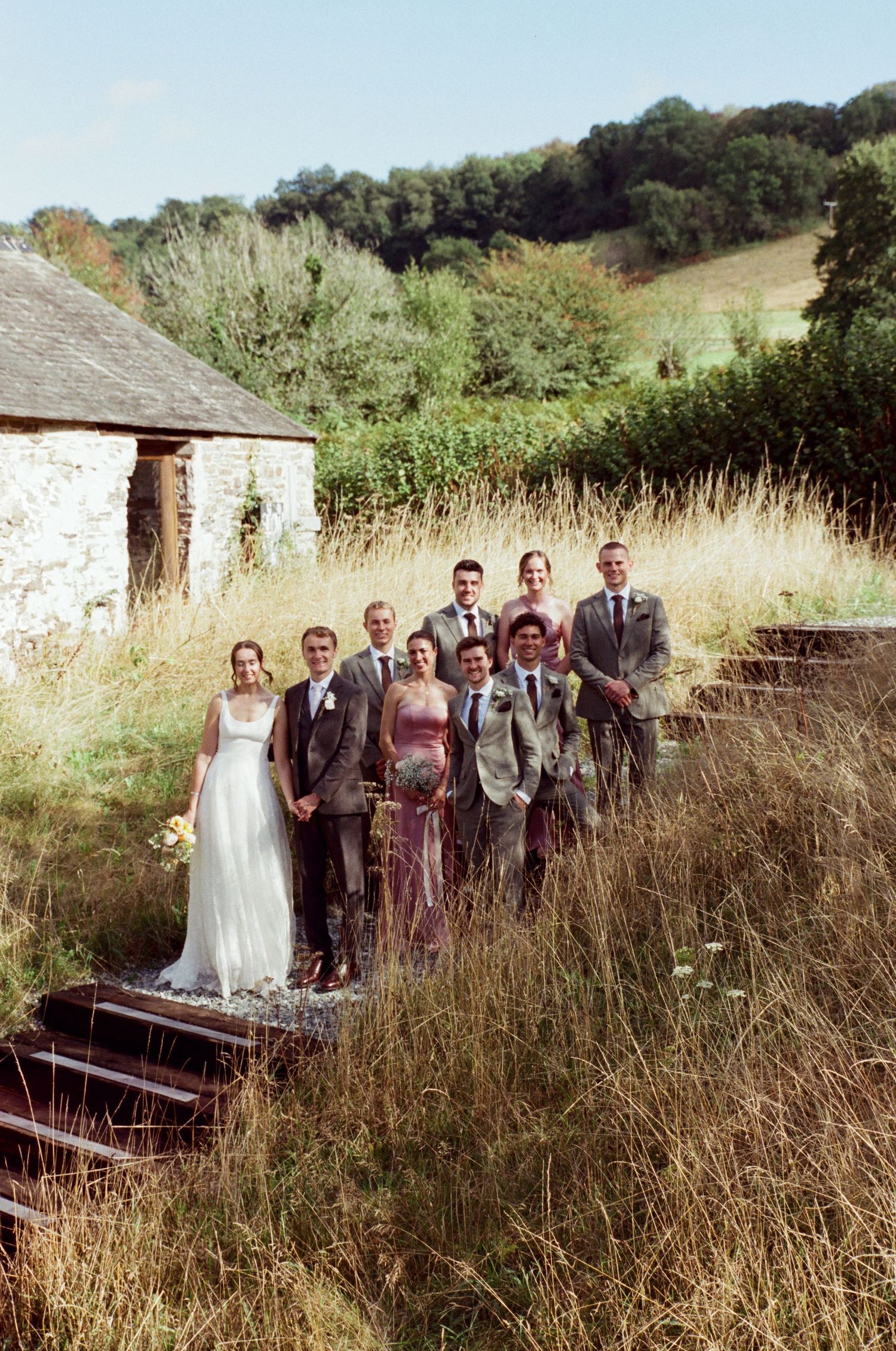 Wedding party poses on rustic stone steps surrounded by wild grass and greenery at a countryside venue.