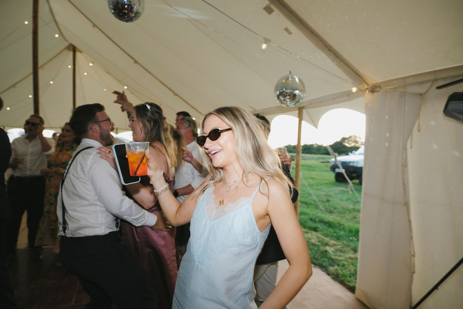 Woman in sunglasses dancing at an outdoor wedding reception tent party with disco balls and guests.