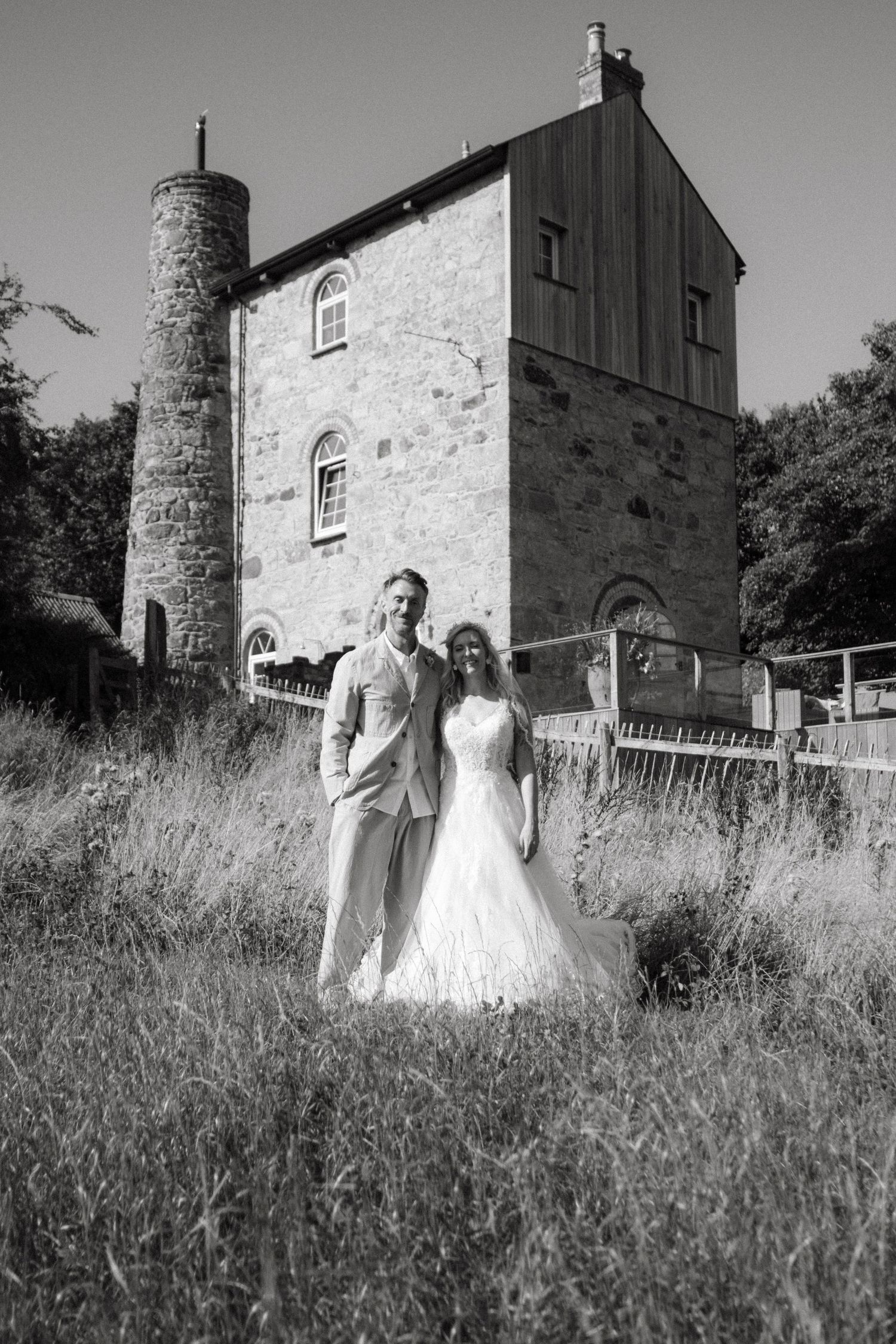 Black and white portrait of bride and groom standing before a rustic stone building with a round tower.