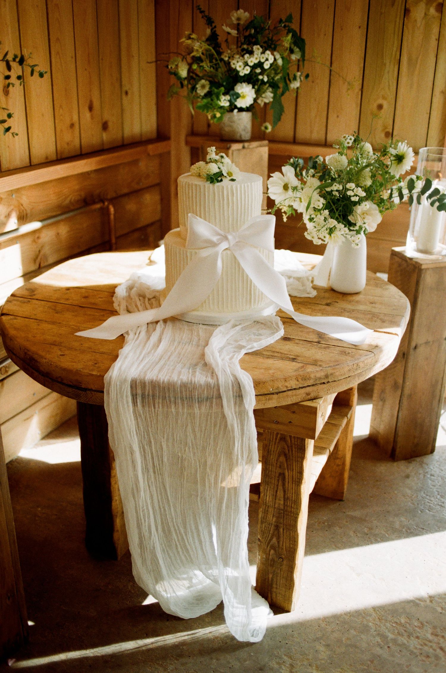 Elegant two-tier white wedding cake with bow on rustic wooden table with white floral arrangements.