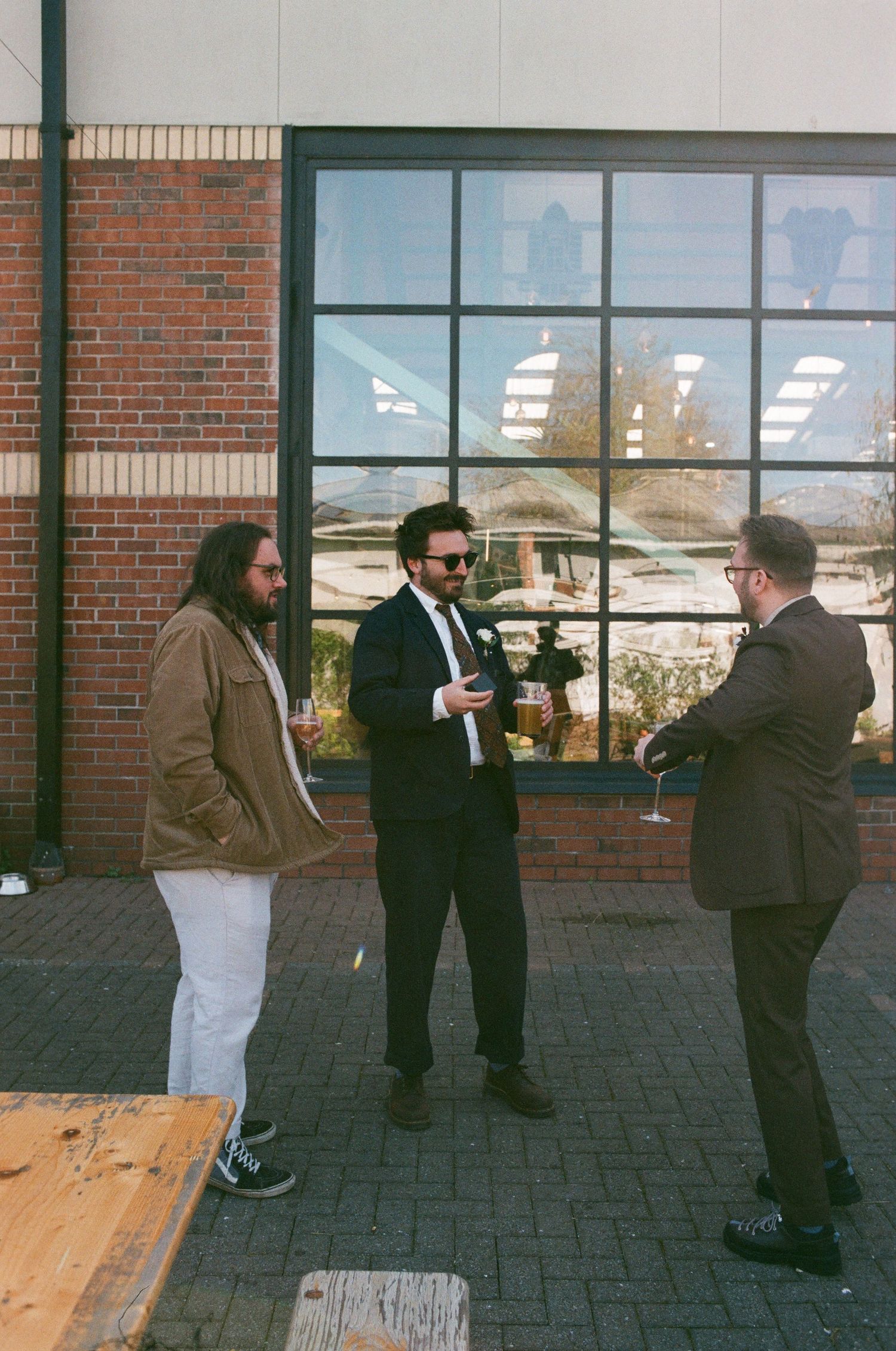 Three men in casual and formal attire conversing outside a brick building with large windows.