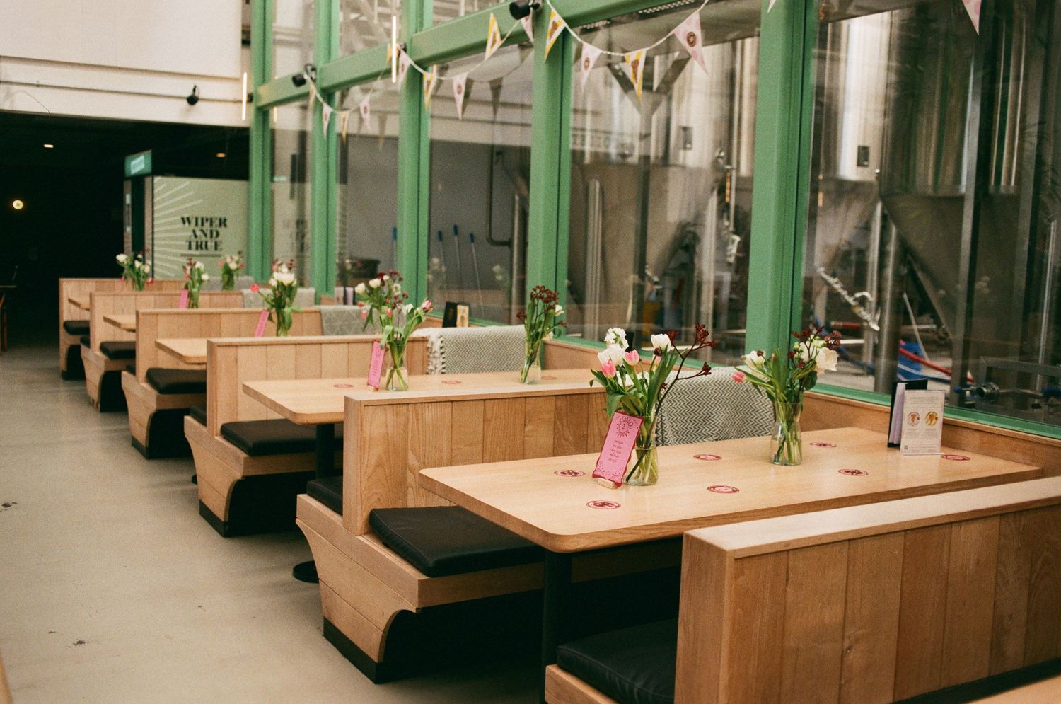 Bright brewery taproom with wooden booth seating, pink tulips in glass vases, and industrial steel tanks visible in the background.