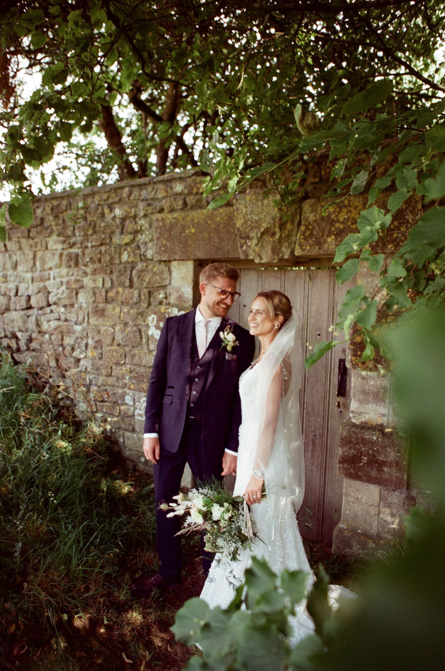 A bride and groom stand together by a rustic stone wall and wooden door, surrounded by lush greenery, holding a white bouquet.