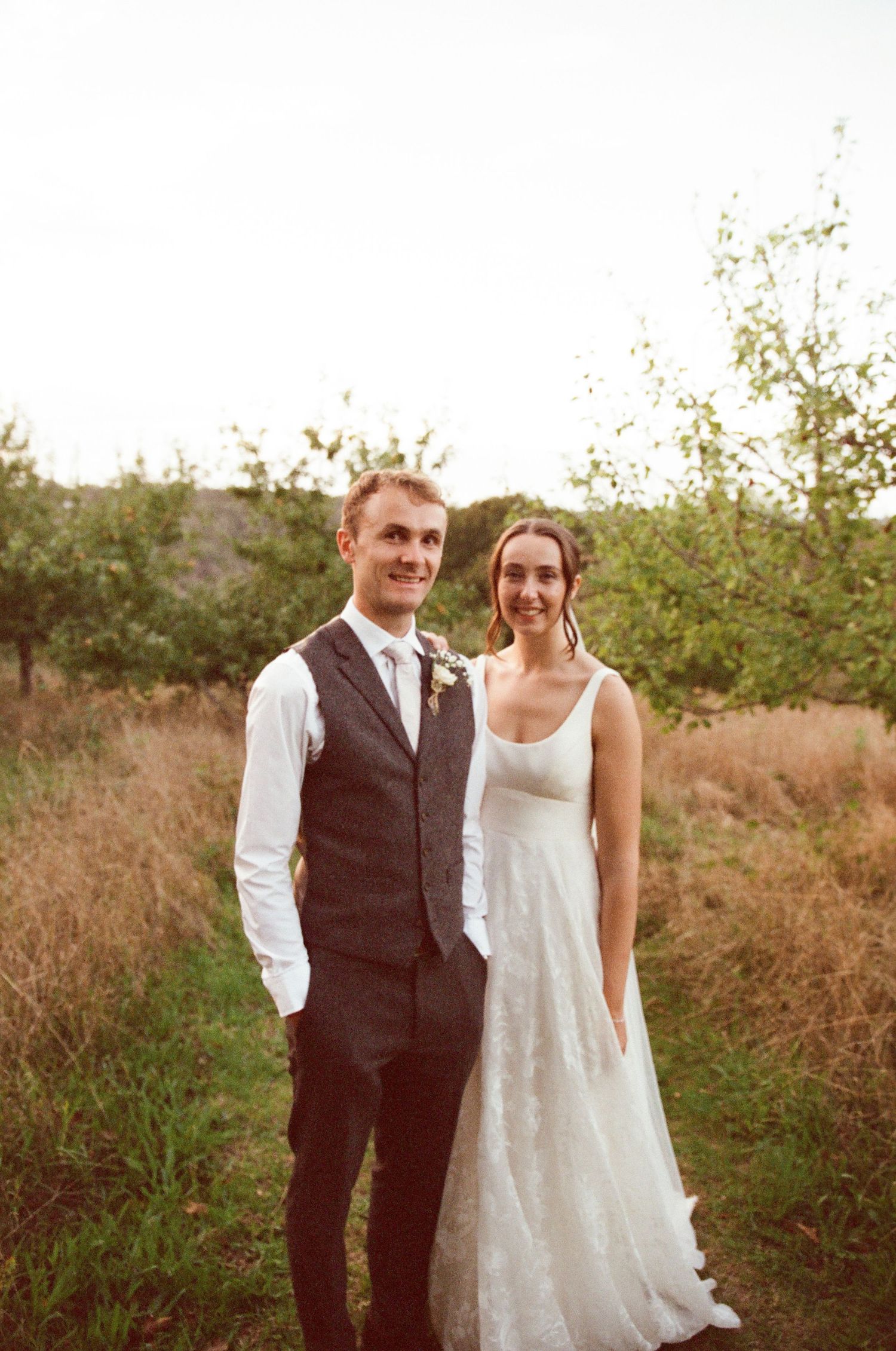 Bride in white lace gown and groom in dark vest and tie pose together outdoors in a golden field at sunset.