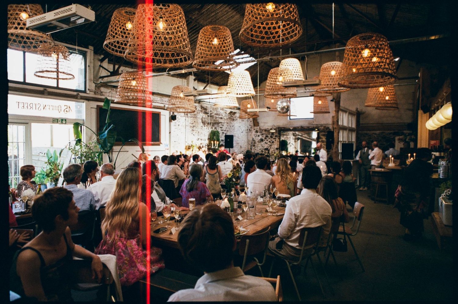 Busy restaurant interior with wicker pendant lights, crowded dining tables, and guests enjoying meals in a modern industrial space.