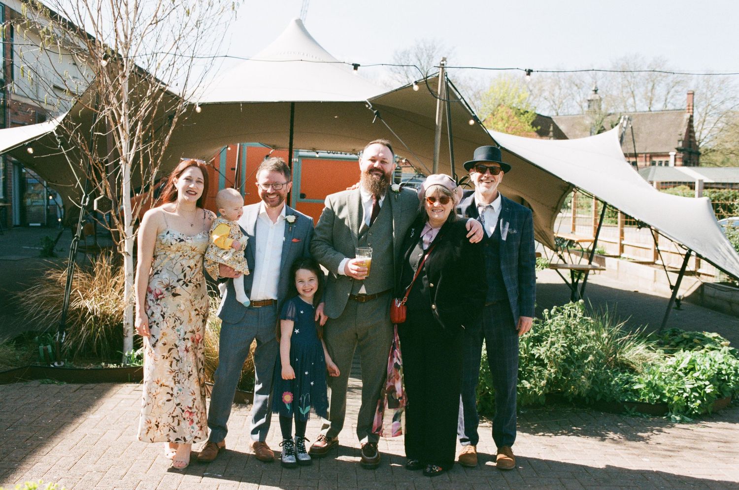 Group of seven people posing outdoors near a large tent at a sunny outdoor event or wedding celebration.