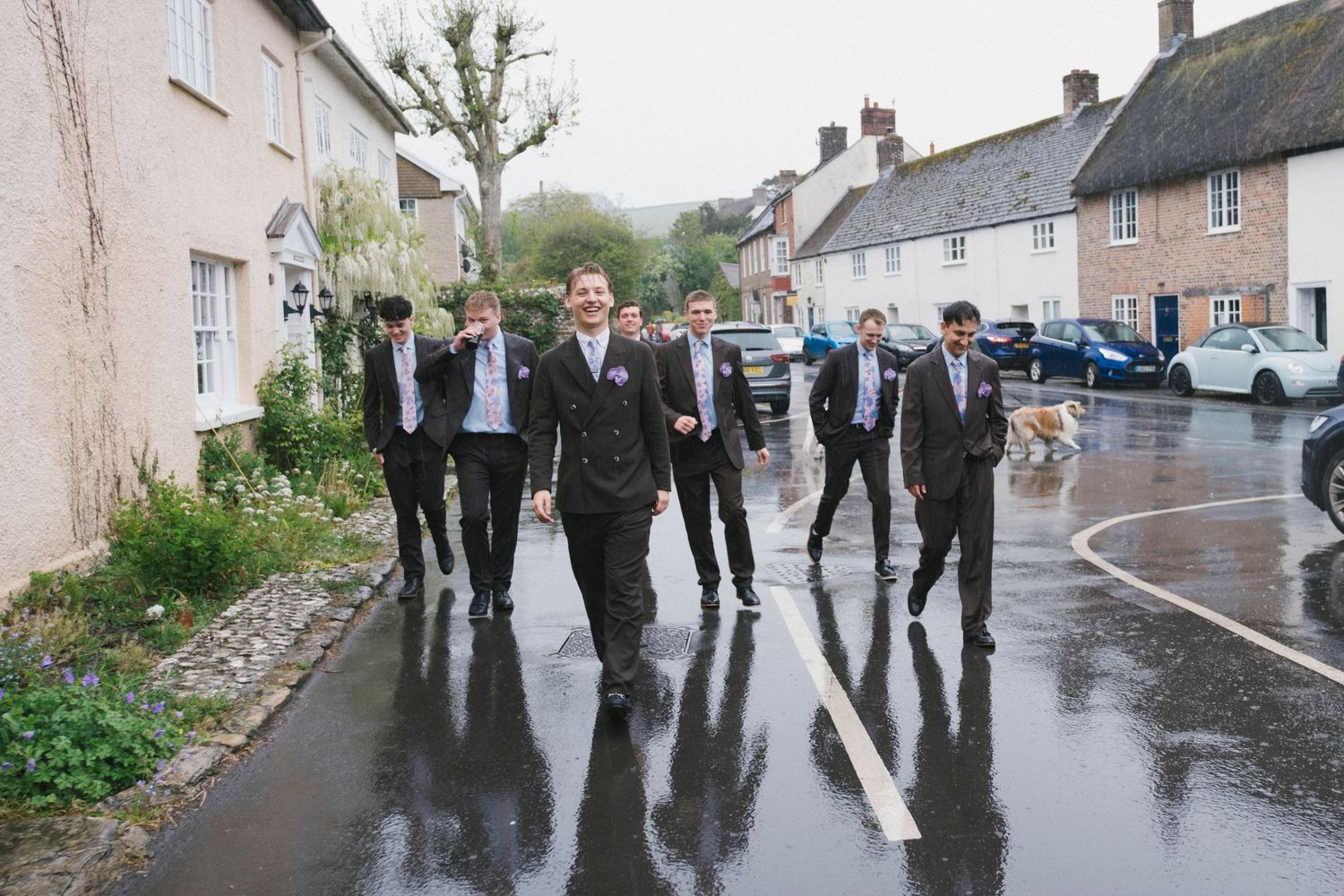 Groom leads groomsmen in dark suits walking confidently down wet village road on wedding day.