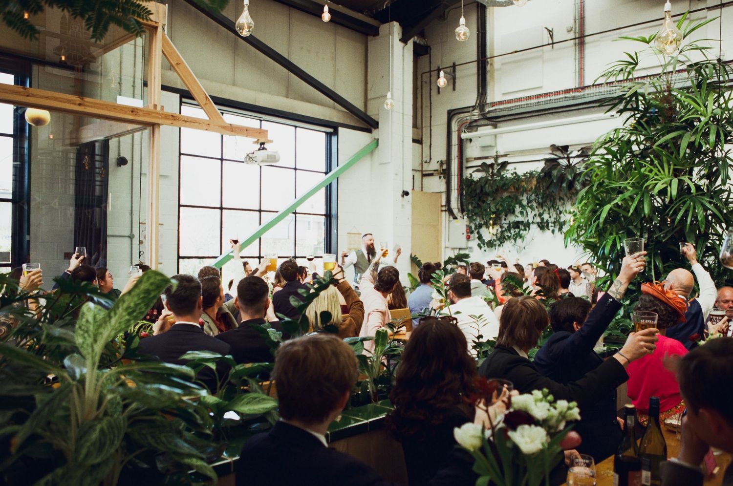 Guests celebrate at an indoor wedding reception surrounded by lush tropical plants in an industrial venue.