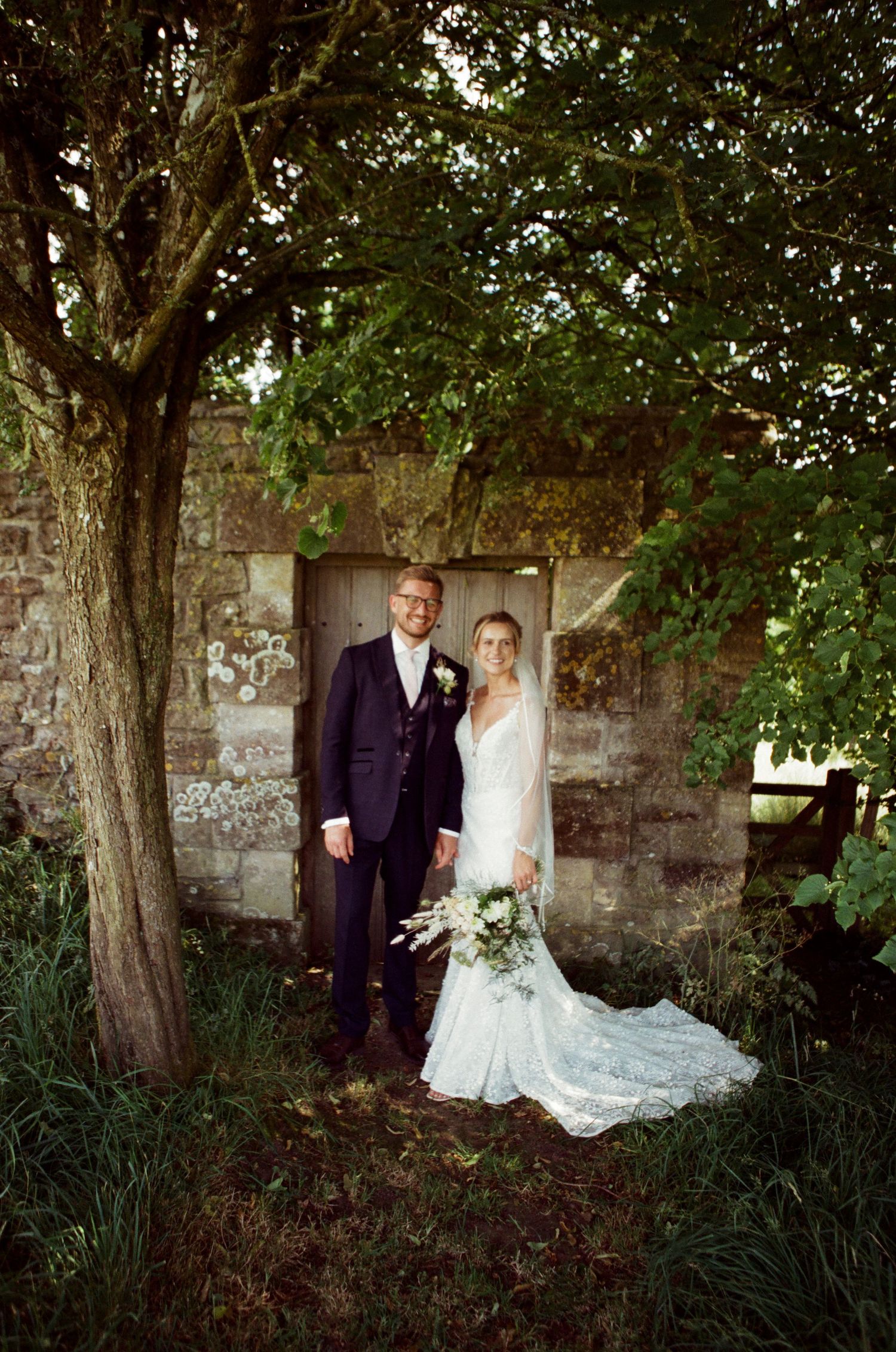 Bride and groom pose under a tree beside a rustic stone building, bride holding a white bouquet in a lace gown.
