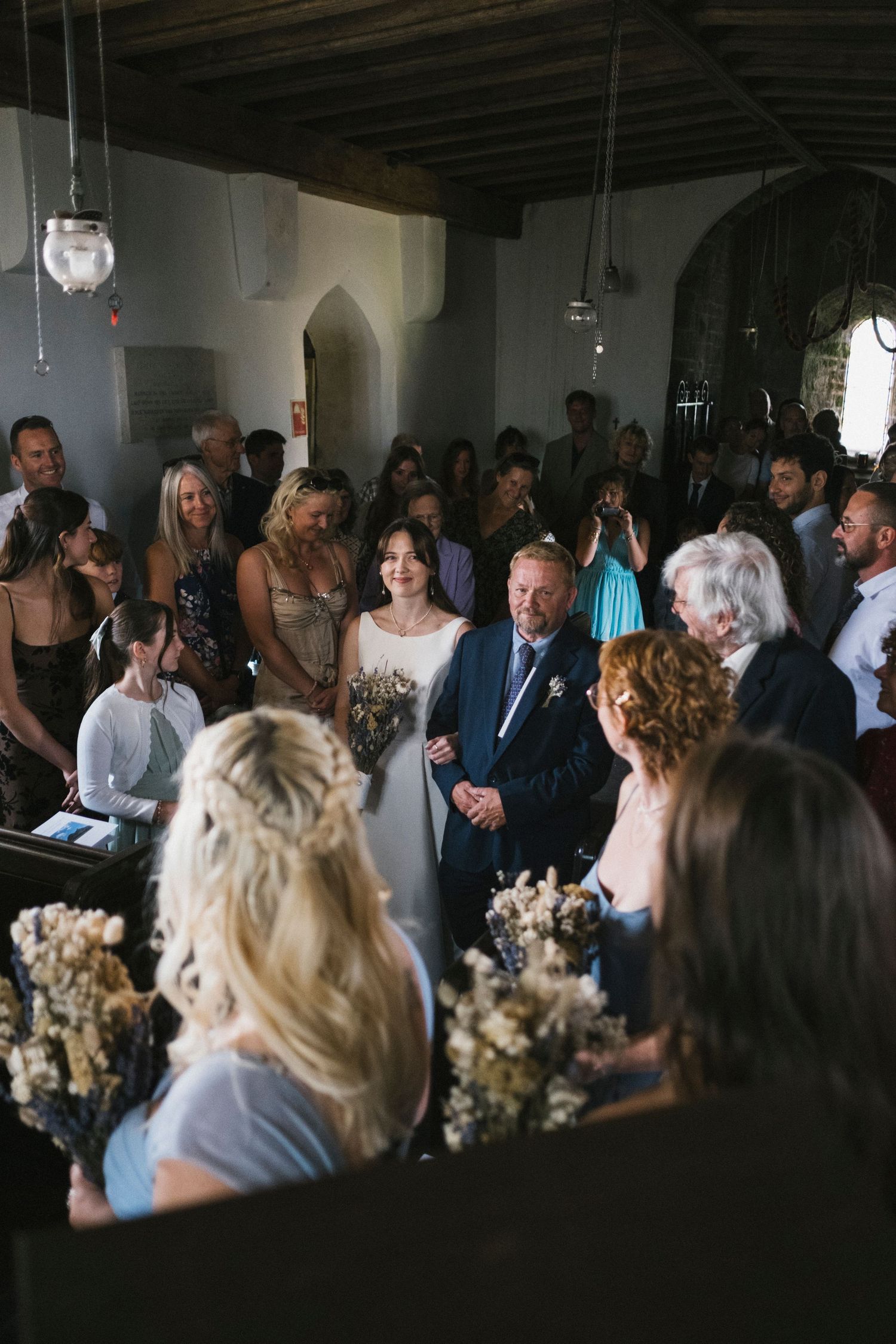 Bride walking down the aisle with her father through a crowd of guests in a rustic stone church.