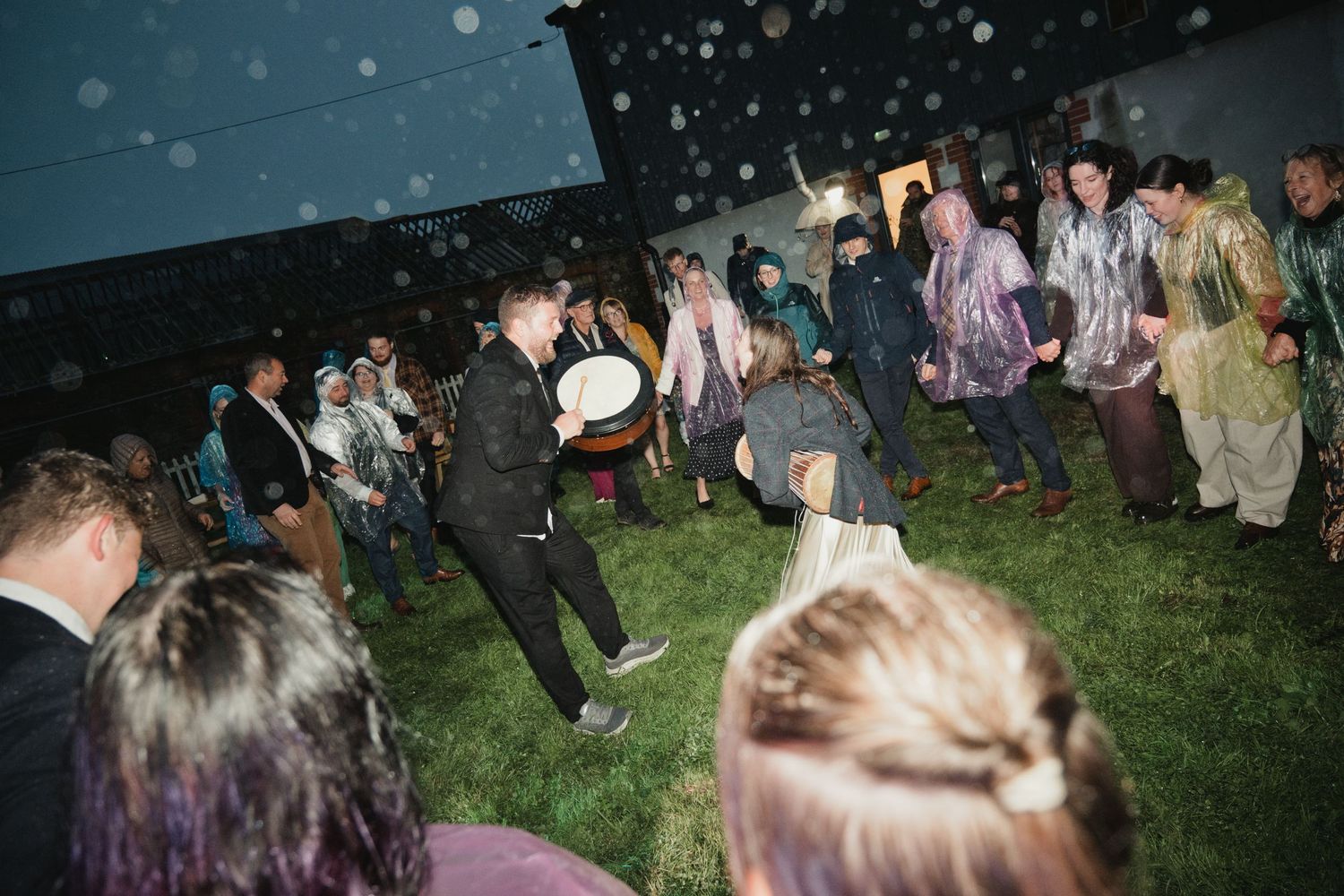 Group of people dancing outdoors at a rainy night event on grass, wearing rain ponchos under dark sky.