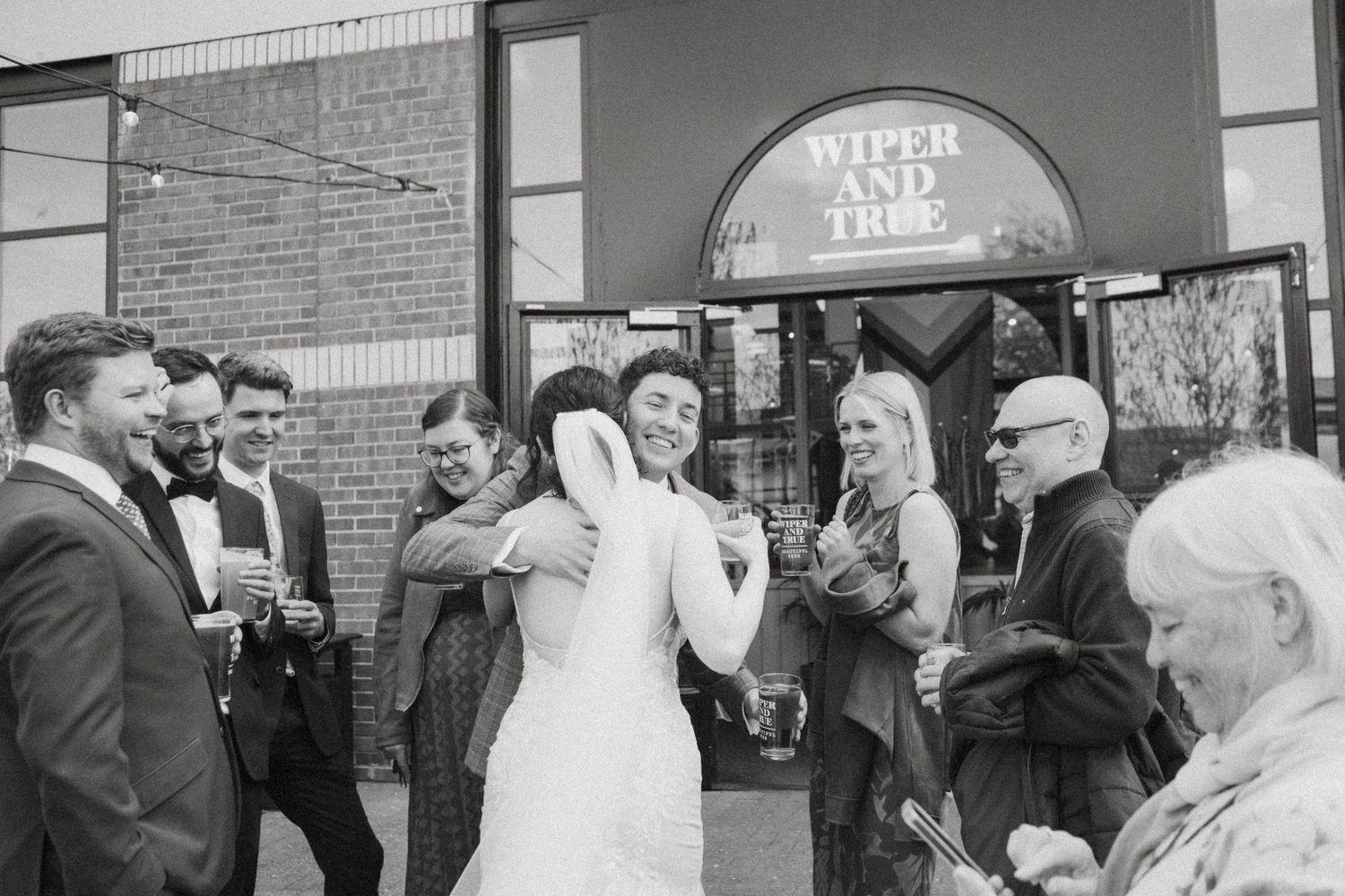 Bride and guest embrace joyfully outside Wiper and True brewery, surrounded by wedding party guests in black and white photo.