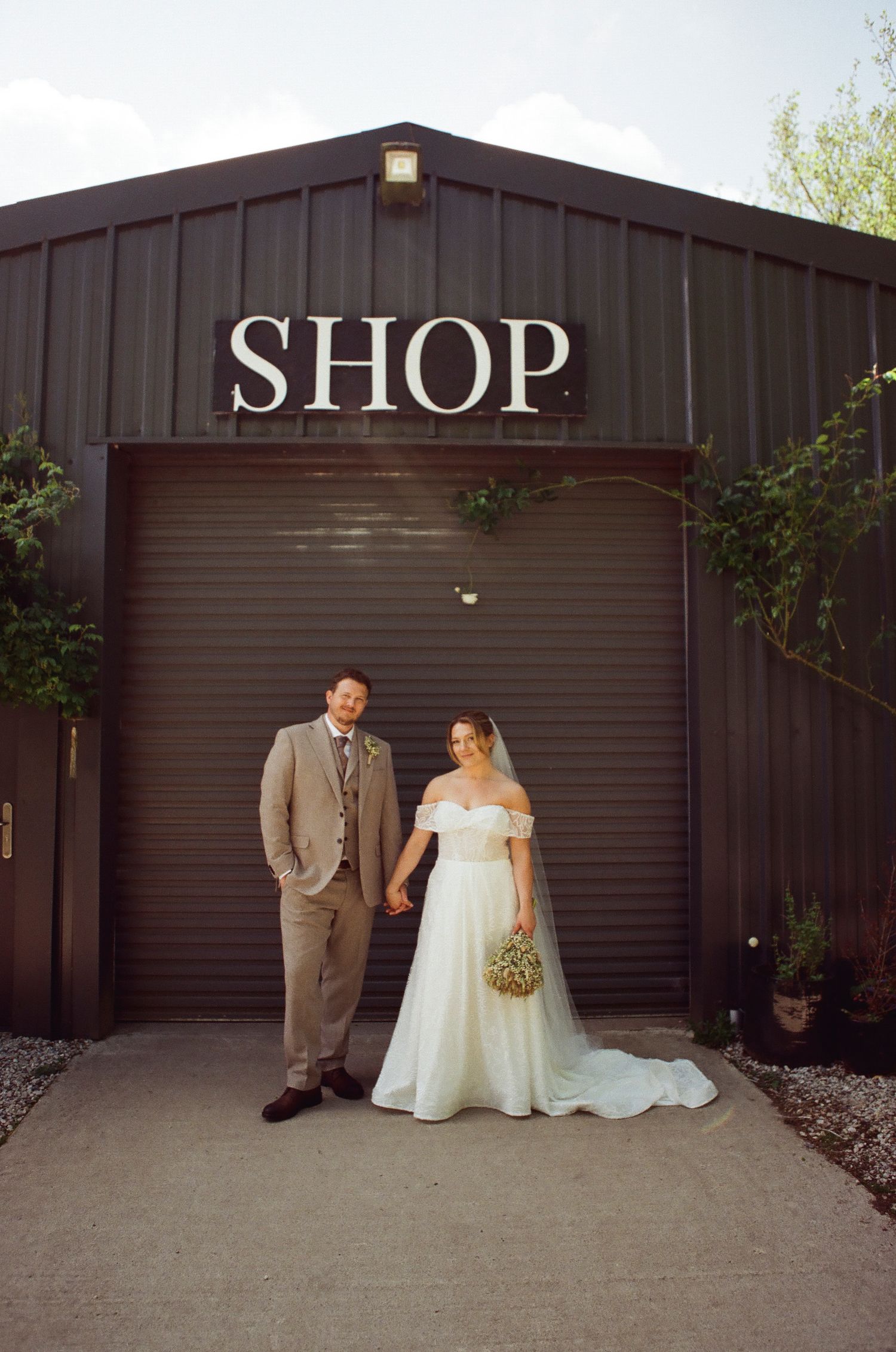 Bride and groom holding hands smiling outside a dark metal shop building on their wedding day.