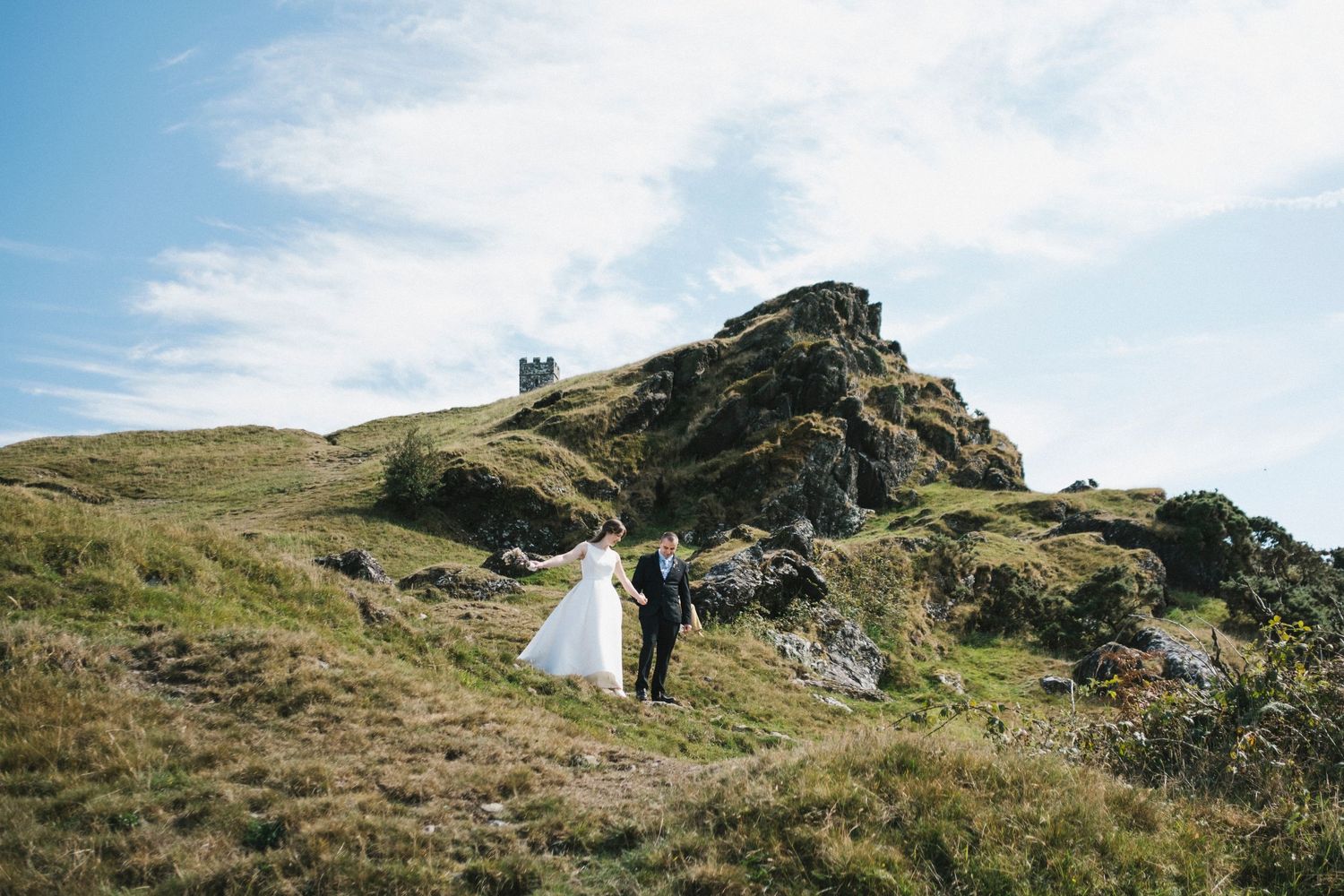 Bride and groom walking hand in hand up scenic green hillside with rocky summit and blue sky.