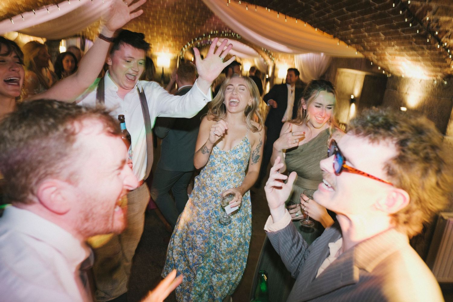 Energetic wedding guests dancing and laughing joyfully under fairy lights at a lively reception celebration.