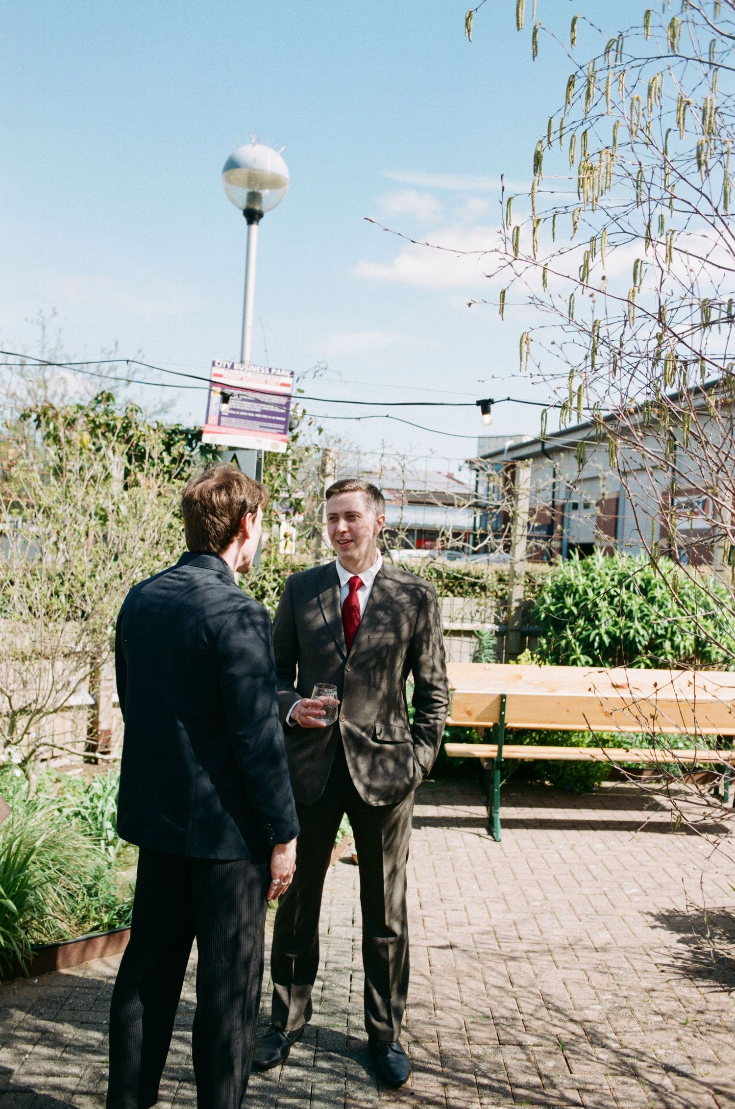 Two men in suits share a conversation outdoors on a sunny day near a park bench and string lights.
