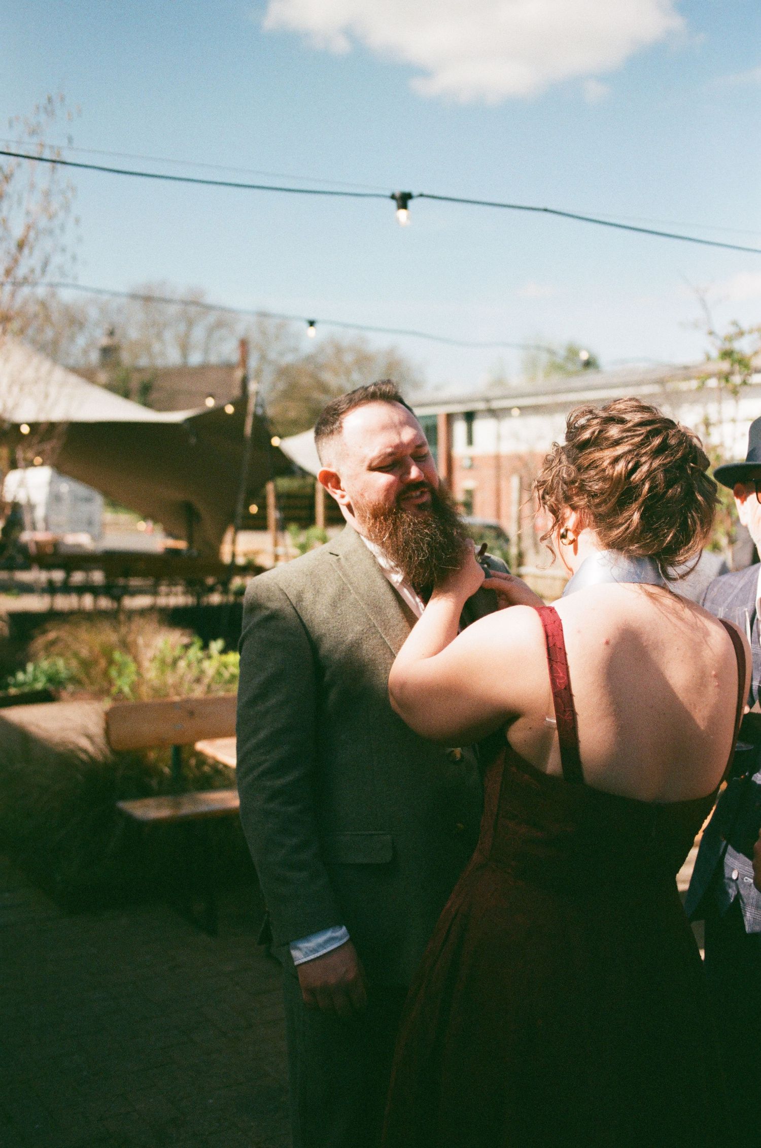 A bearded man in a suit shares a tender moment with a woman in a red dress at an outdoor wedding reception.