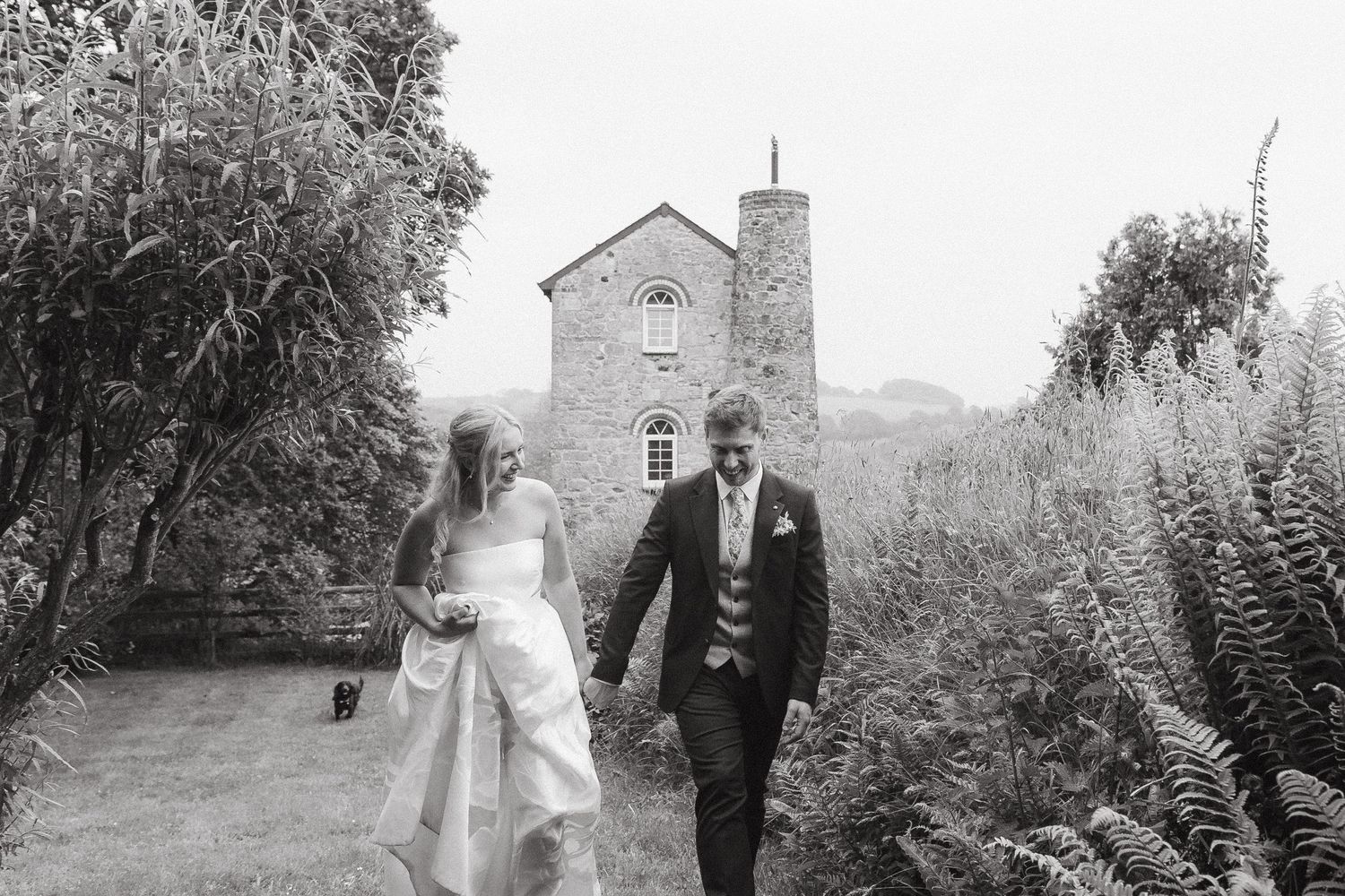 Bride and groom walk hand in hand through a garden path beside a historic stone chapel on their wedding day.