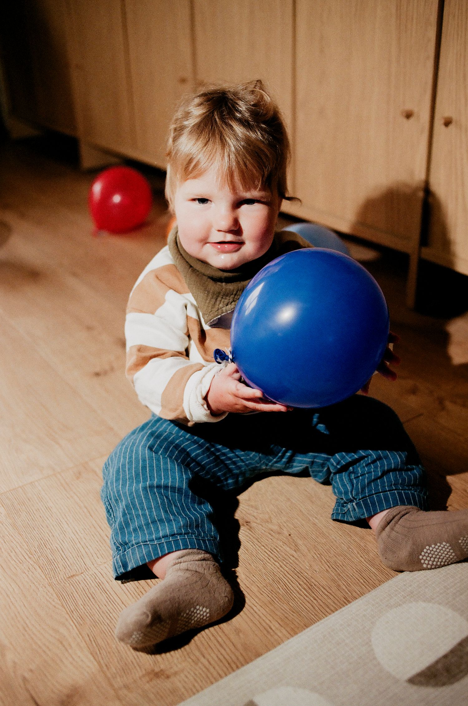 Cute toddler sitting on floor holding a blue balloon with a red balloon behind.