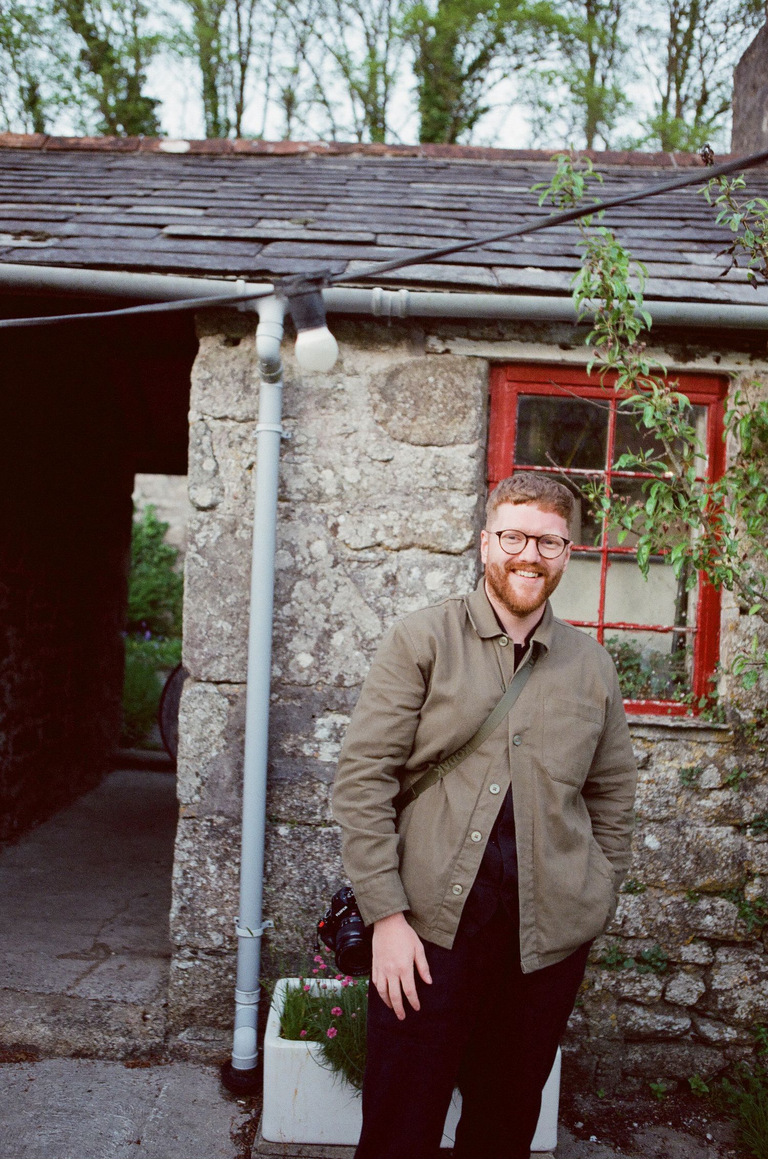 A smiling man in a green jacket stands outside a rustic stone cottage with a red window.