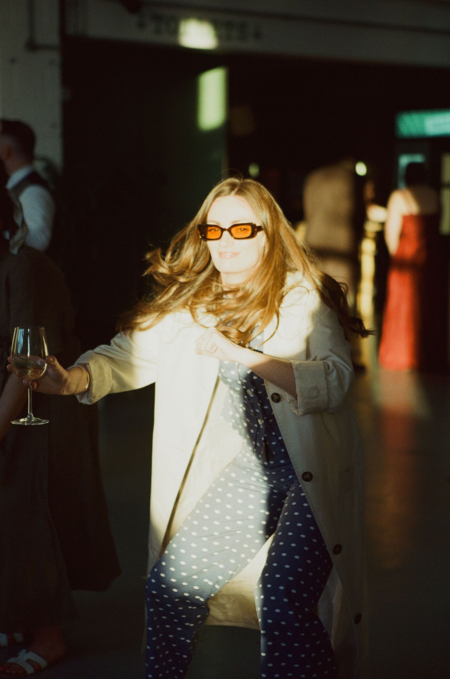 Stylish wedding guest with long blonde hair wearing orange sunglasses holds a champagne glass at an evening reception.