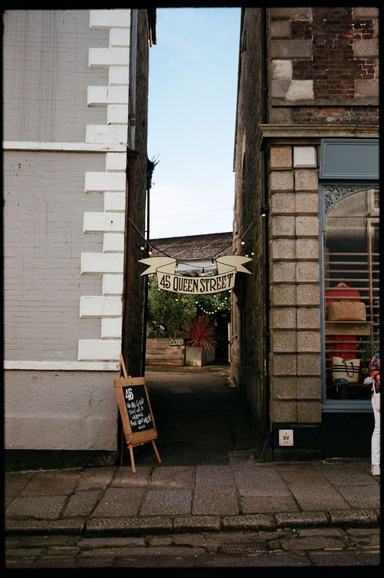 Narrow alleyway entrance to 45 Queen Street with a chalkboard sign and string lights between historic stone buildings.