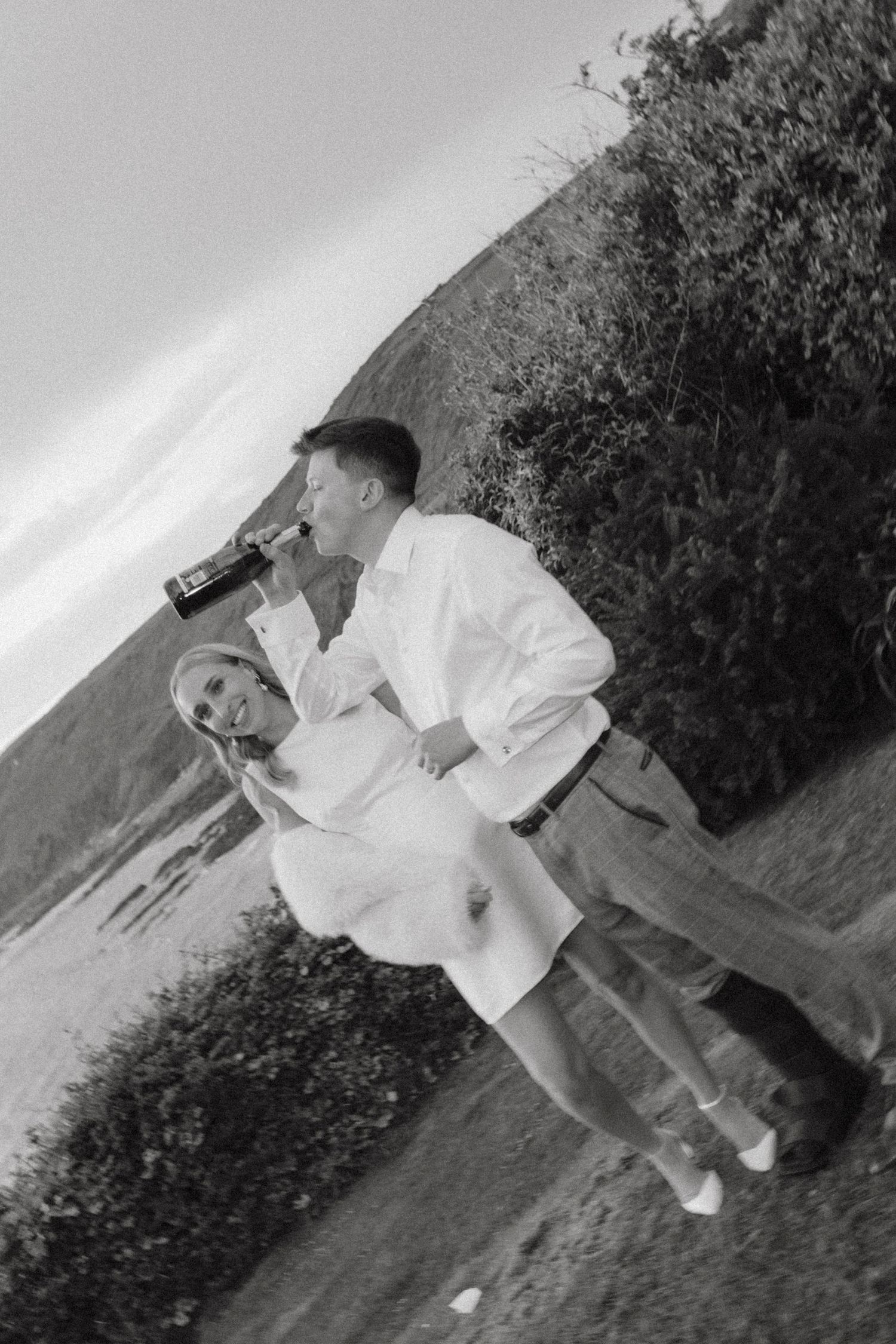 Black and white photo of a couple outdoors, man drinking from bottle while woman in white dress smiles at camera.