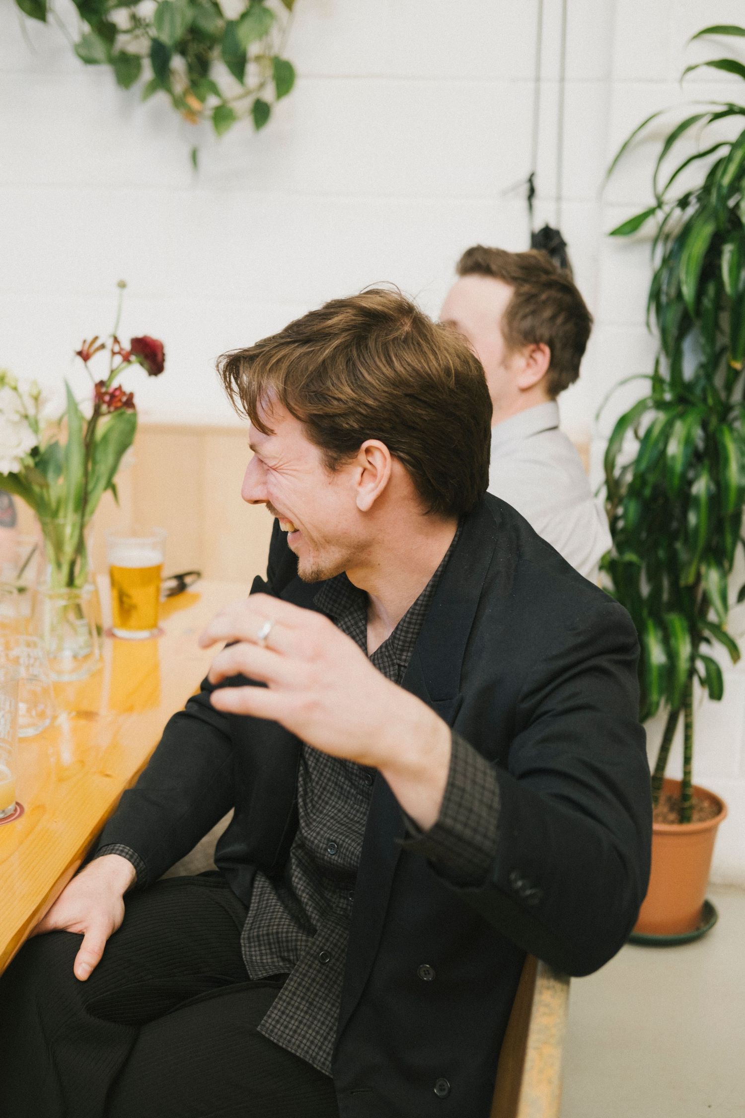 A man in a black blazer laughs at a table with beer glasses and plants nearby.