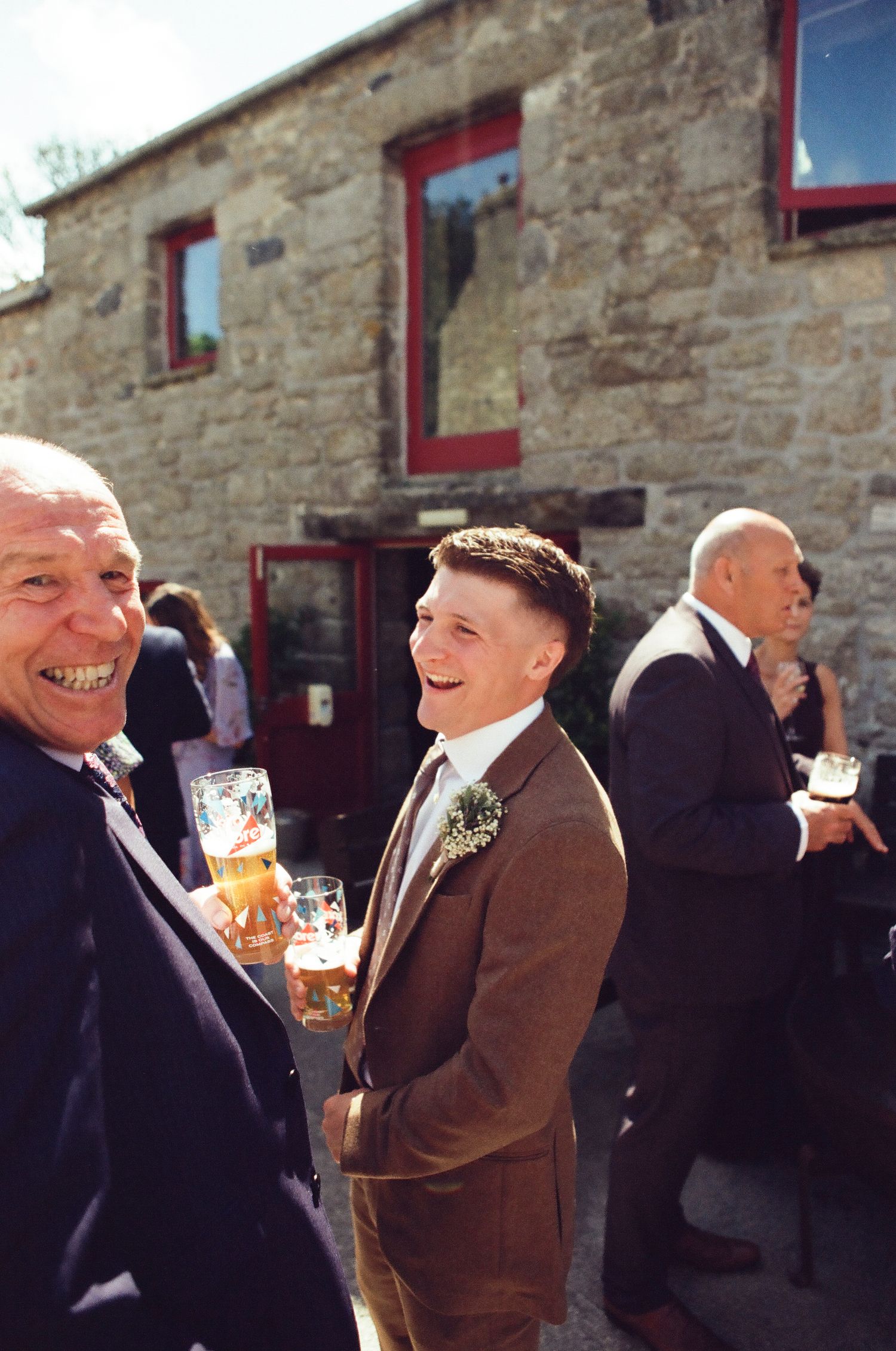 Smiling groom in brown suit holding a beer with guests outside a rustic stone wedding venue.
