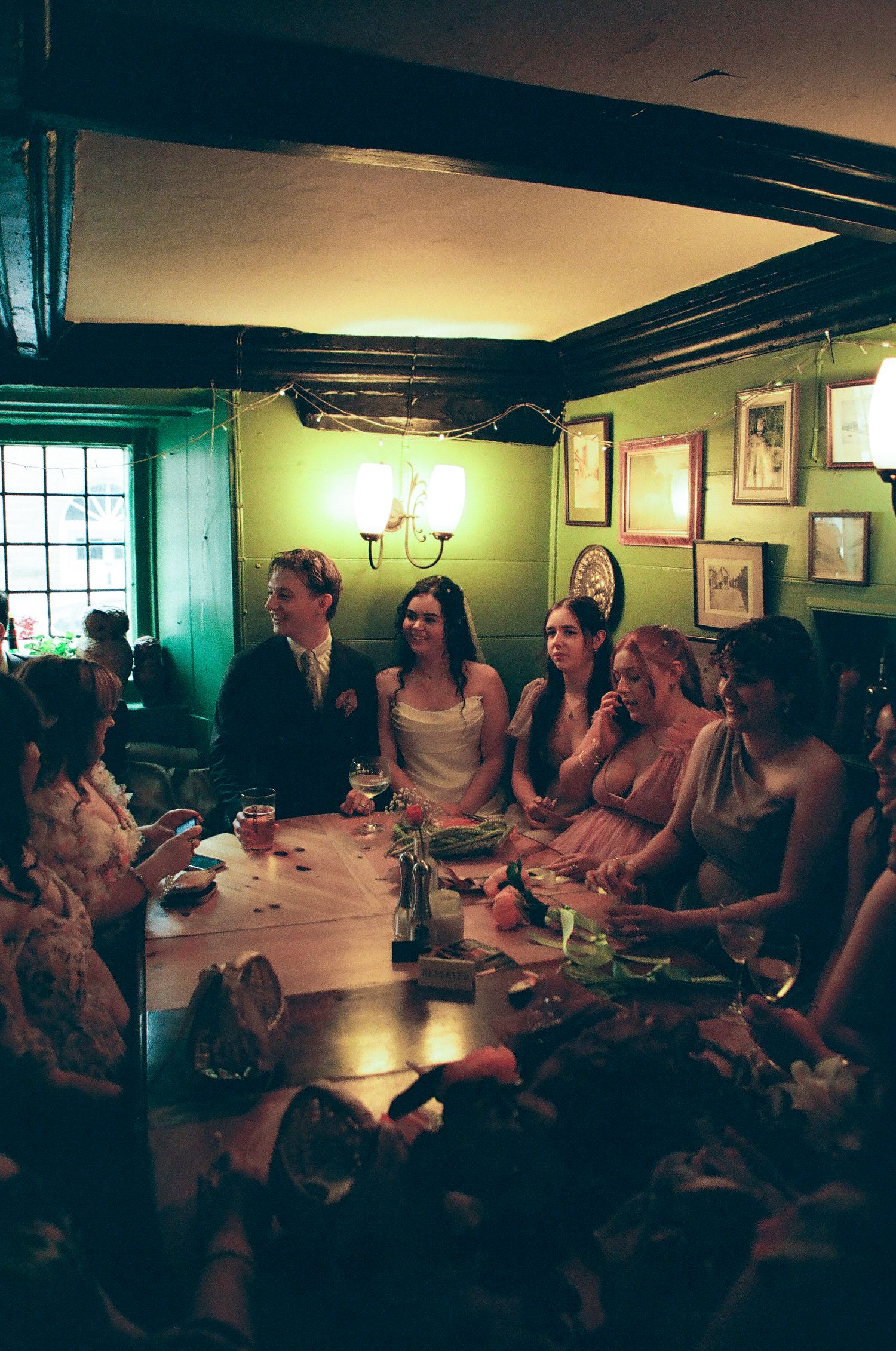 Wedding party gathers around a table in a cozy green-walled pub during the reception.