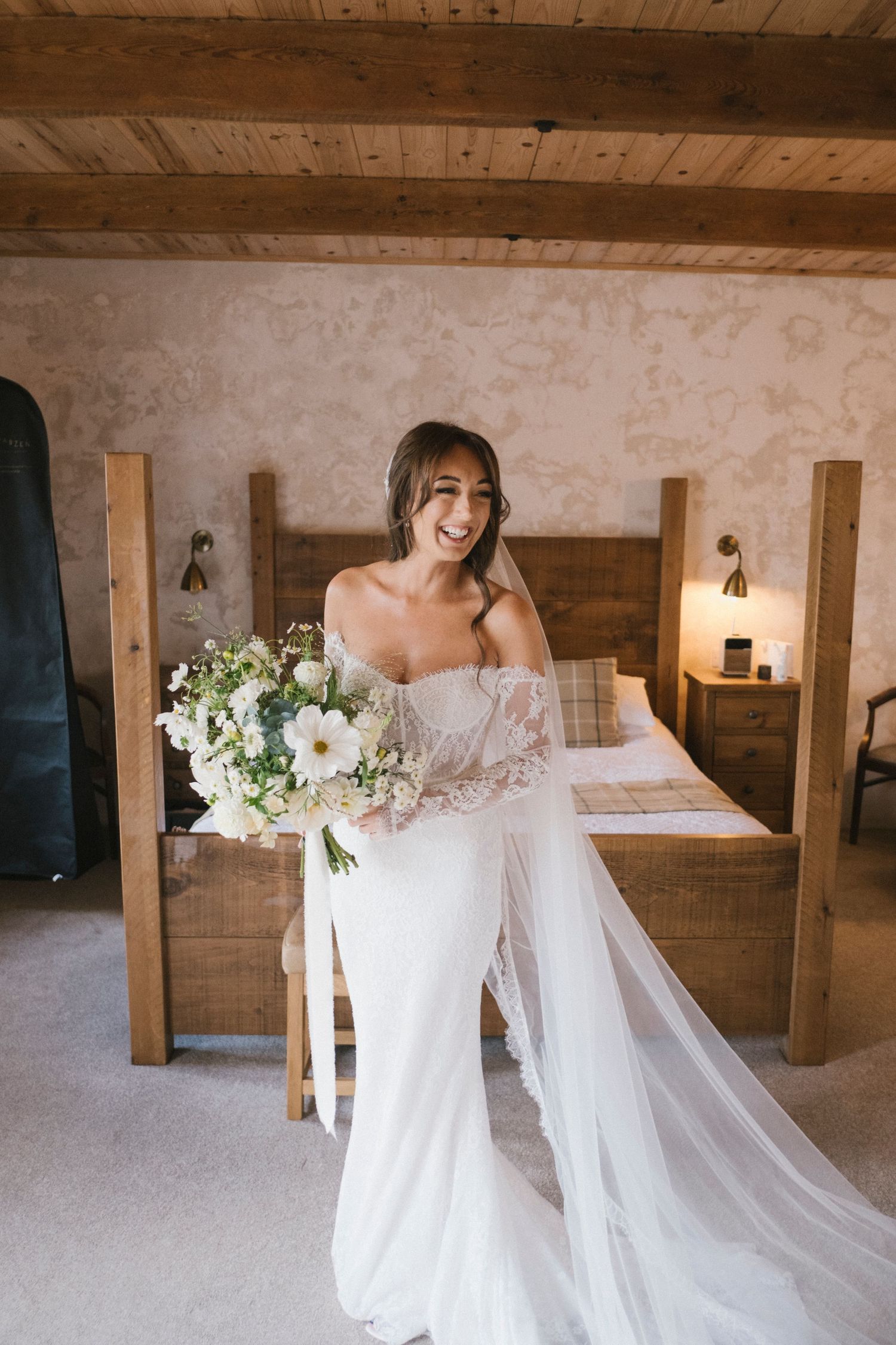 Radiant bride in an off-shoulder gown holds a white floral bouquet in a cozy wooden-beamed bedroom.