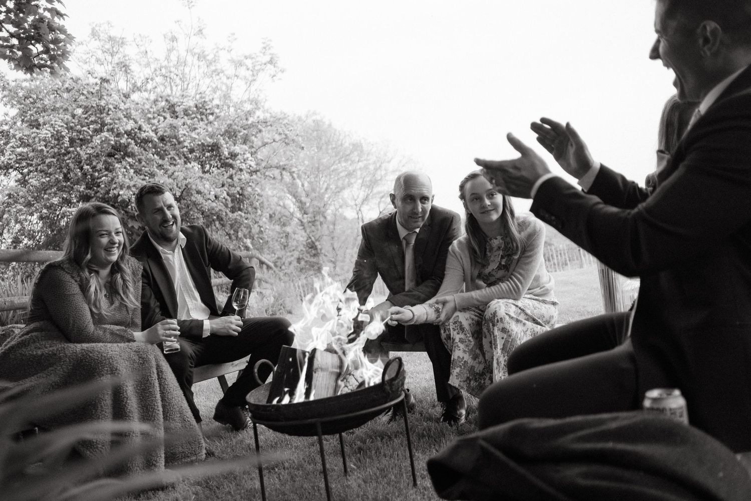 Wedding guests gathered around outdoor fire pit in black and white candid evening photograph.