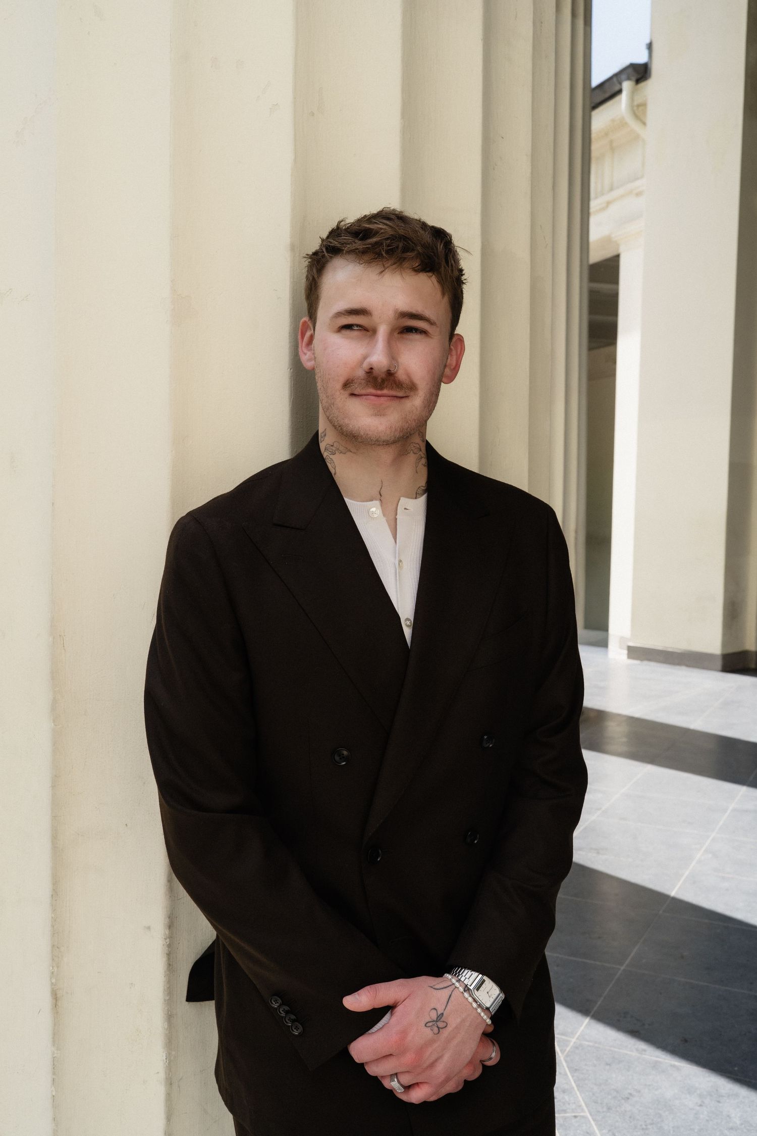 Young man with mustache in black blazer smiling, leaning against white columns in elegant architectural setting.