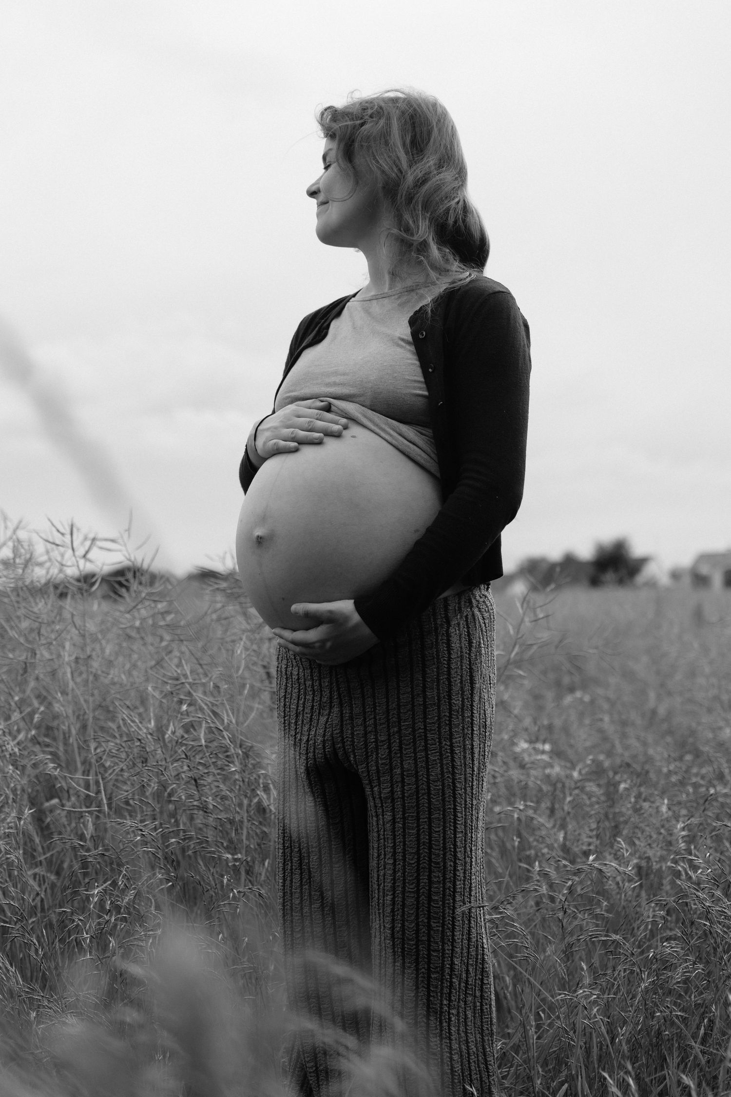 Black and white side profile of pregnant woman holding baby bump in outdoor field setting.