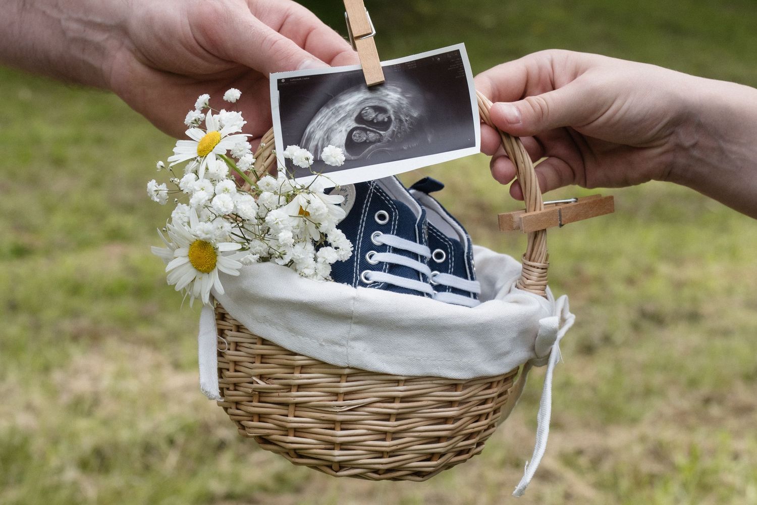 Two hands holding an ultrasound scan above a wicker basket containing baby sneakers and white daisies.