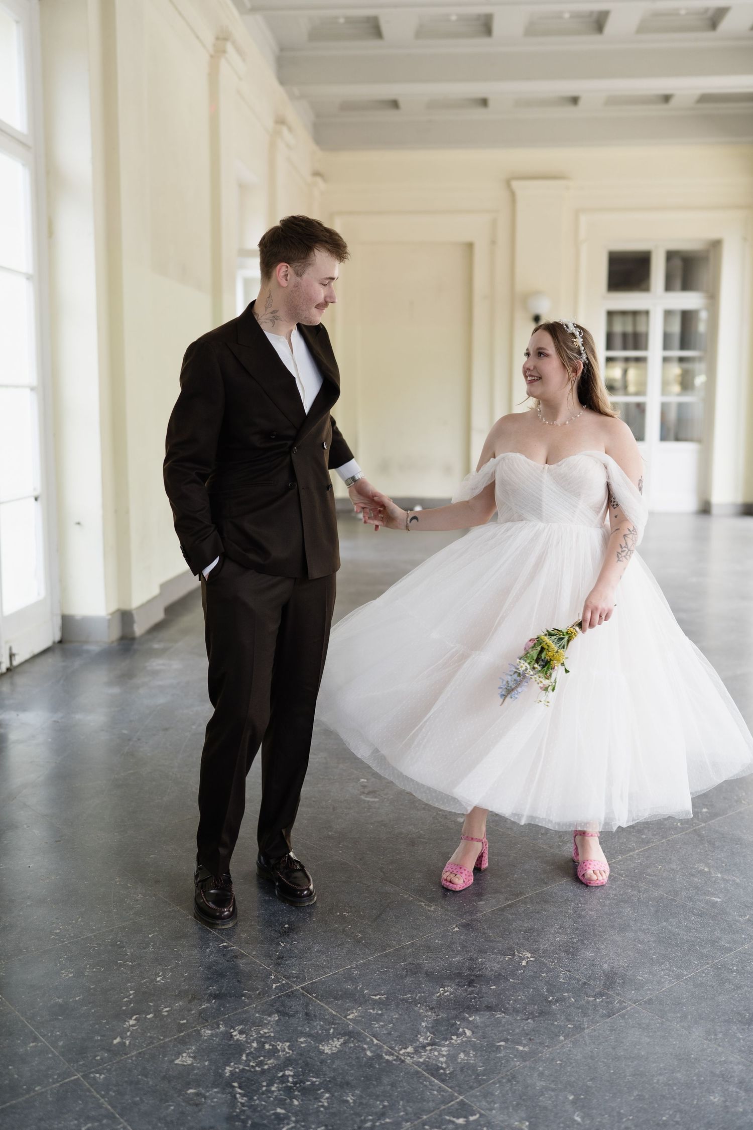 Bride in white tea-length dress with pink heels holds groom's hand while walking in elegant hall.