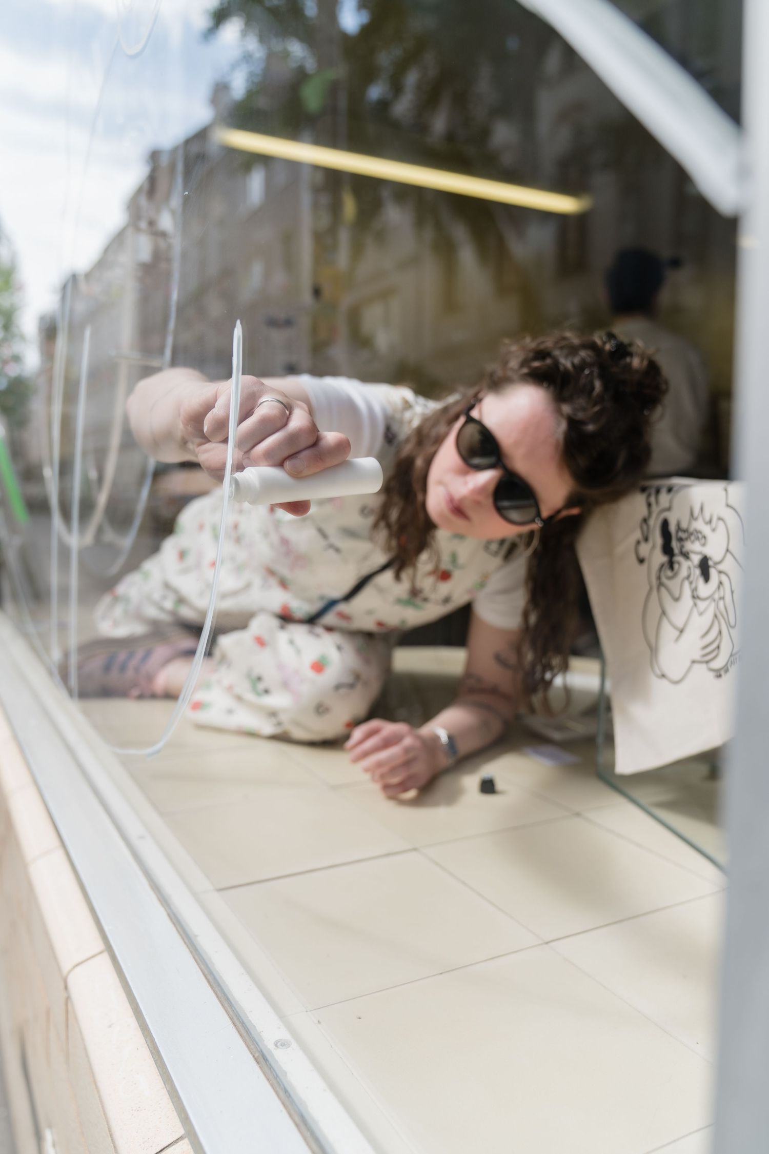 Woman in sunglasses drawing on interior cafe window from floor level.