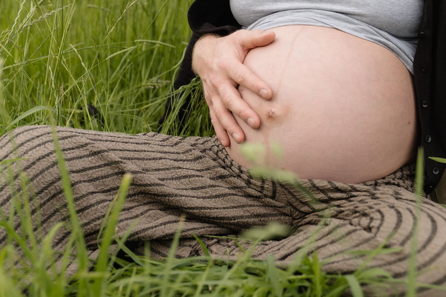Close-up of pregnant woman cradling bare baby bump while sitting outdoors in lush green grass.