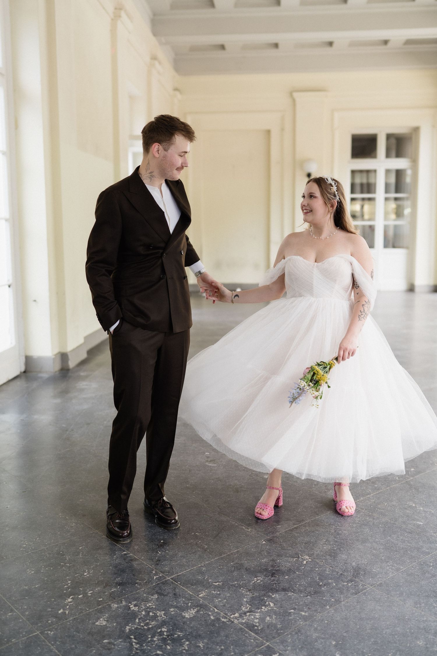 Bride in white tea-length dress with pink heels holds groom's hand in dark suit, walking and smiling indoors.