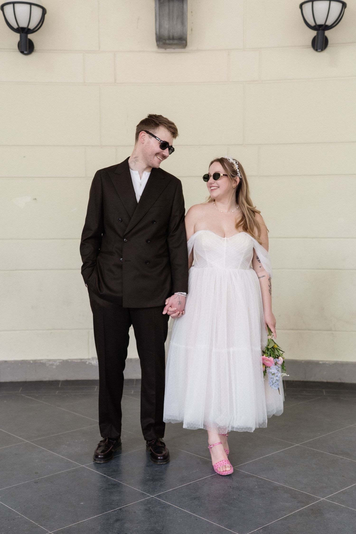 Couple in wedding attire holding hands, groom in dark suit, bride in white tulle dress with pink heels and bouquet.