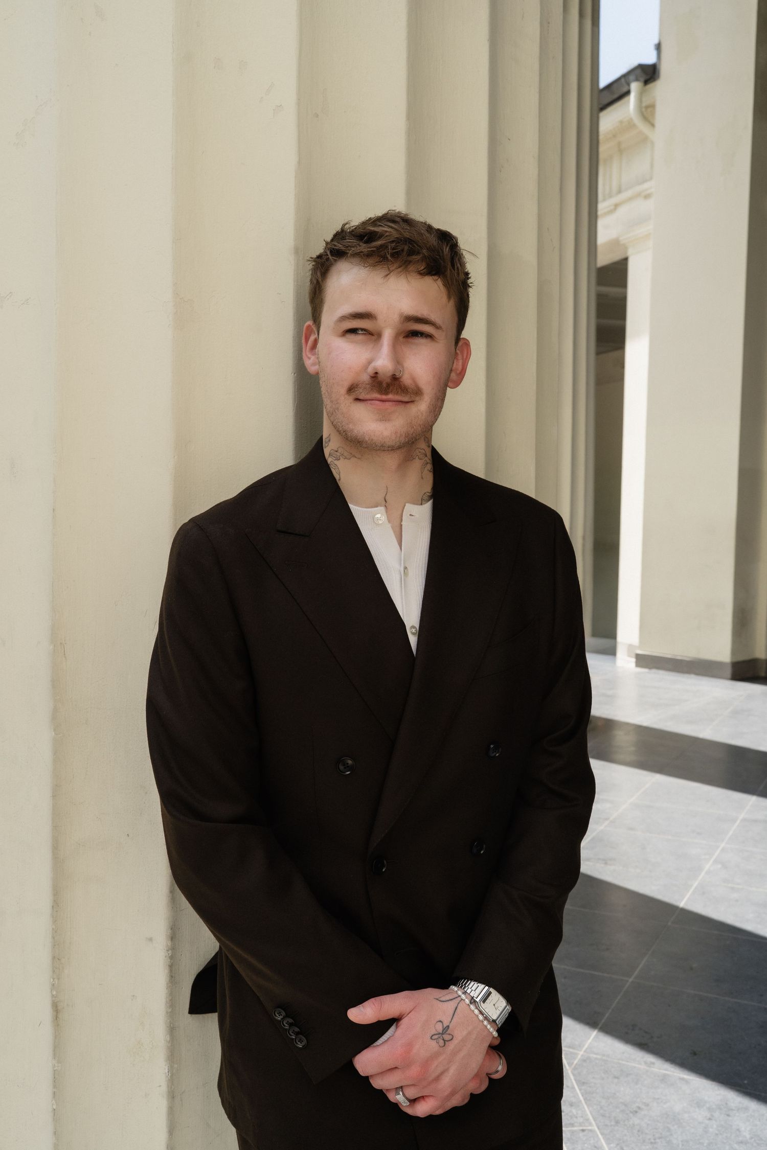 Handsome young groom with mustache smiling in dark double-breasted suit leaning against stone column.