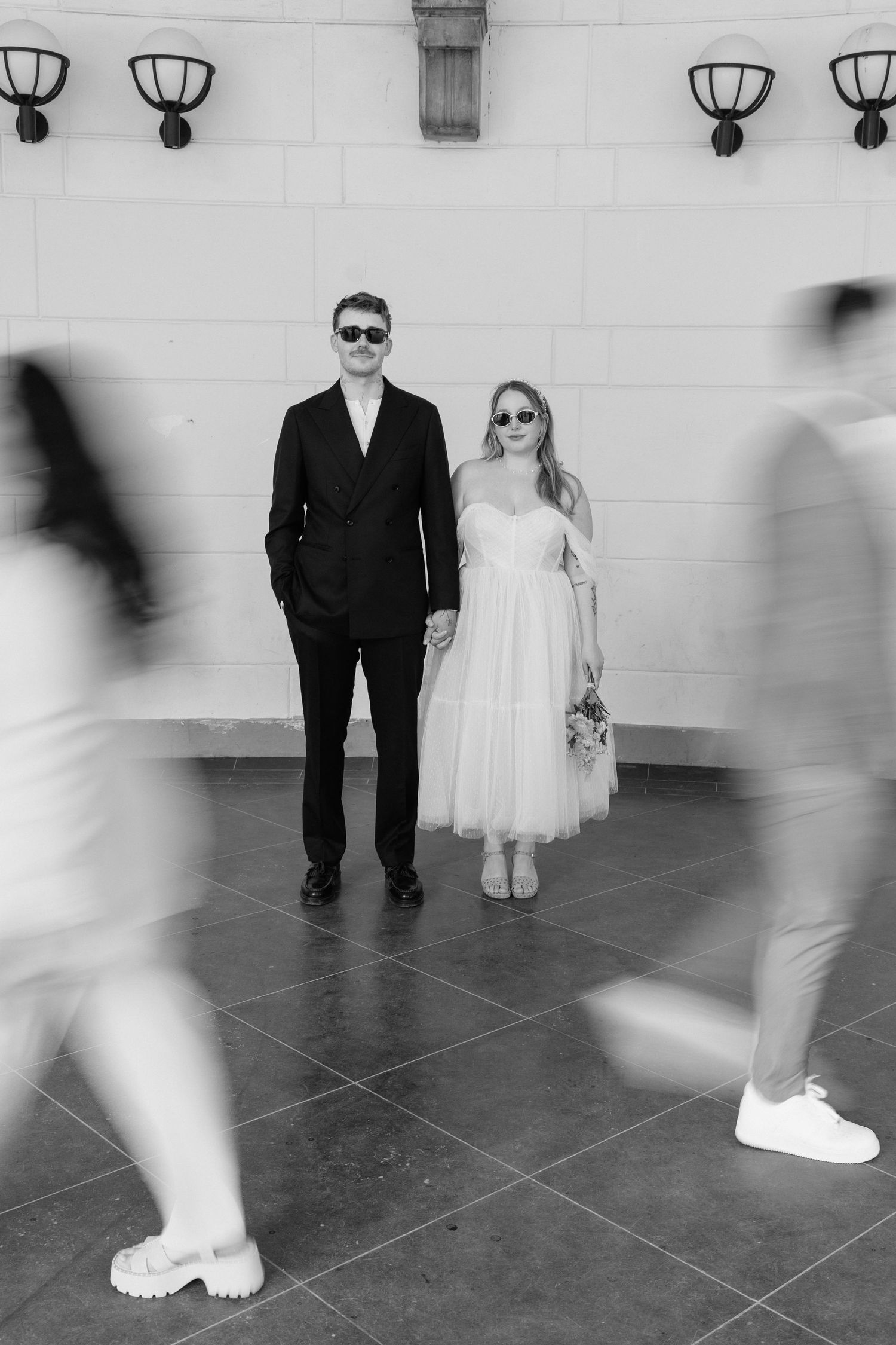 Bride and groom in sunglasses holding hands as blurred guests walk past them in black and white photo.