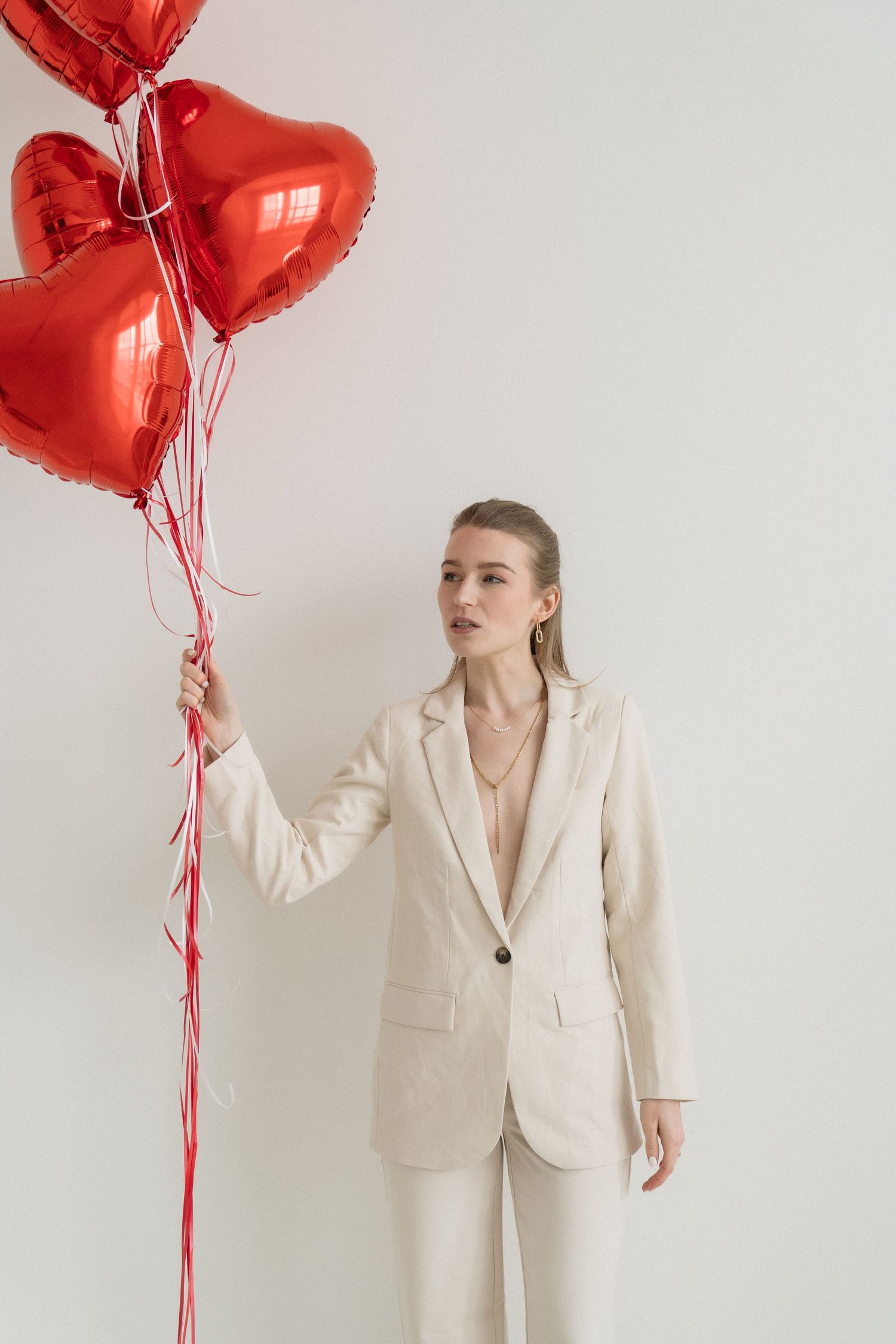 Woman in white suit holding red heart-shaped balloons against a light background.