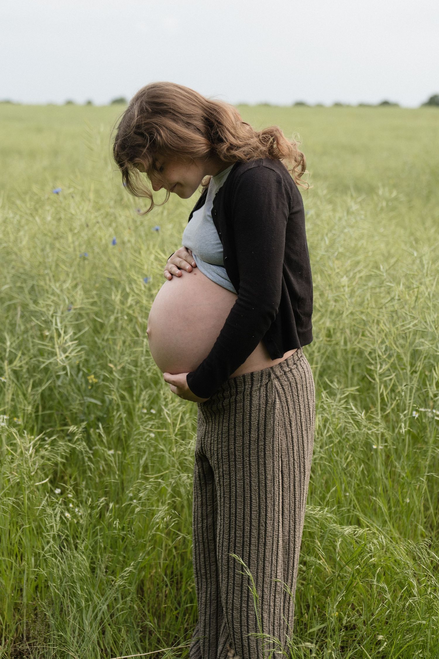 Pregnant woman in a green field cradling her bare baby bump, wearing striped pants and a black cardigan.
