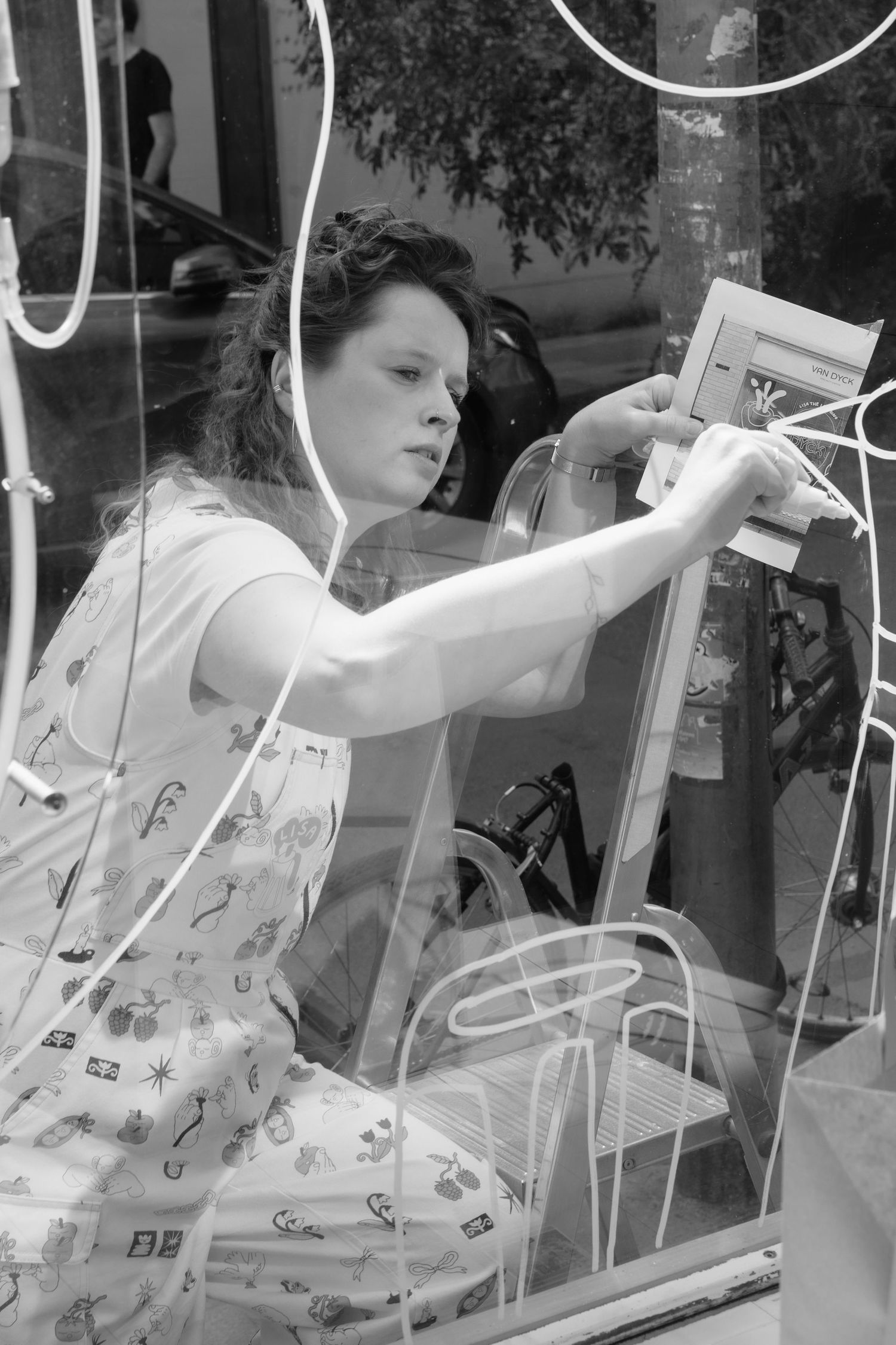 Woman in patterned dress arranging framed artwork in cafe display window.