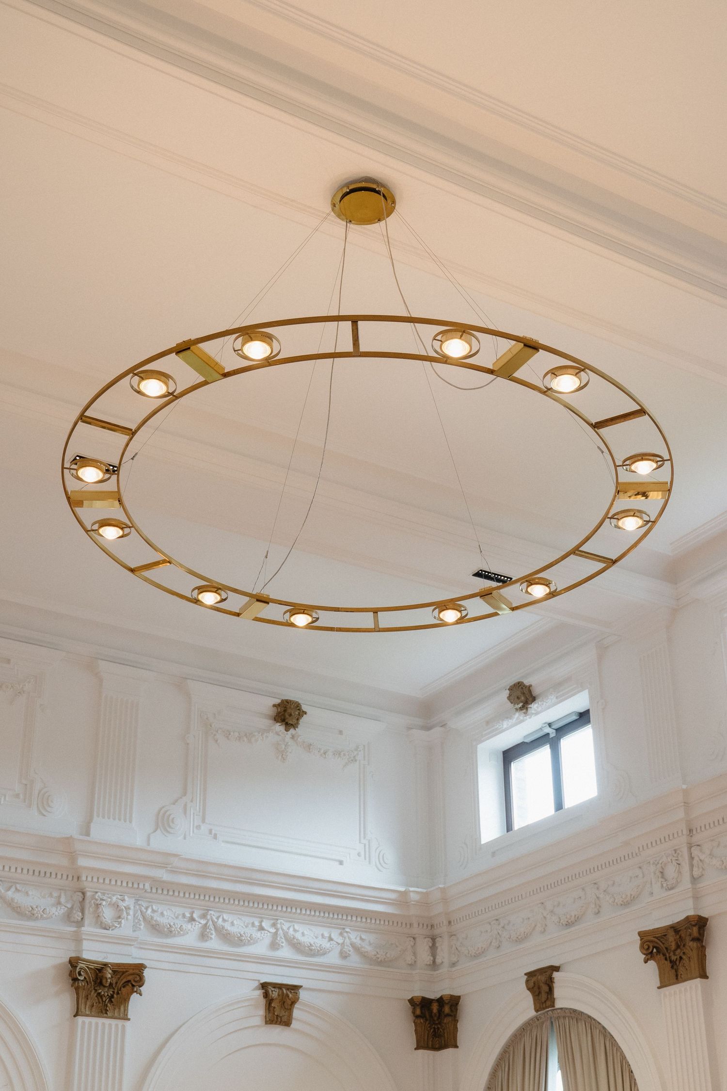 Modern circular gold chandelier with spotlights hanging in a white ornate classical interior.