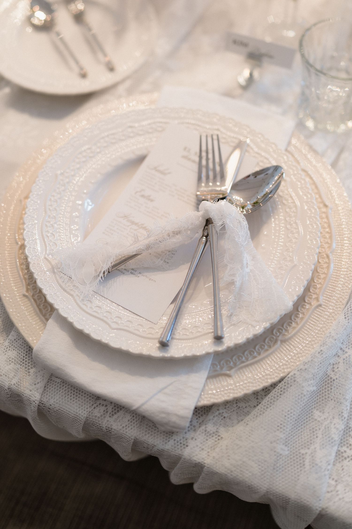 Elegant white wedding place setting with lace charger plate, silver cutlery, and folded napkin.