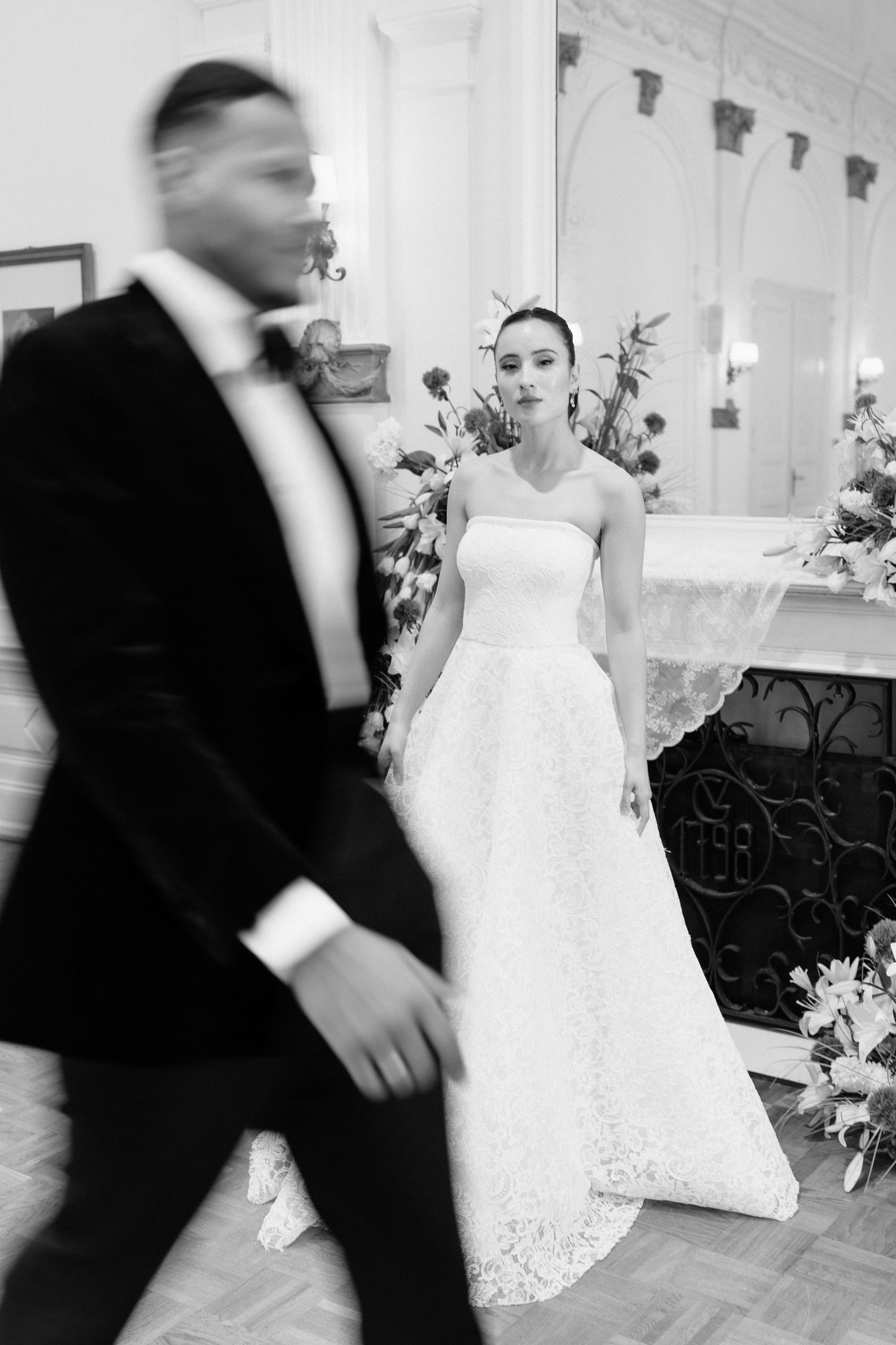 Black and white photo of bride in strapless lace gown as groom walks past in tuxedo.