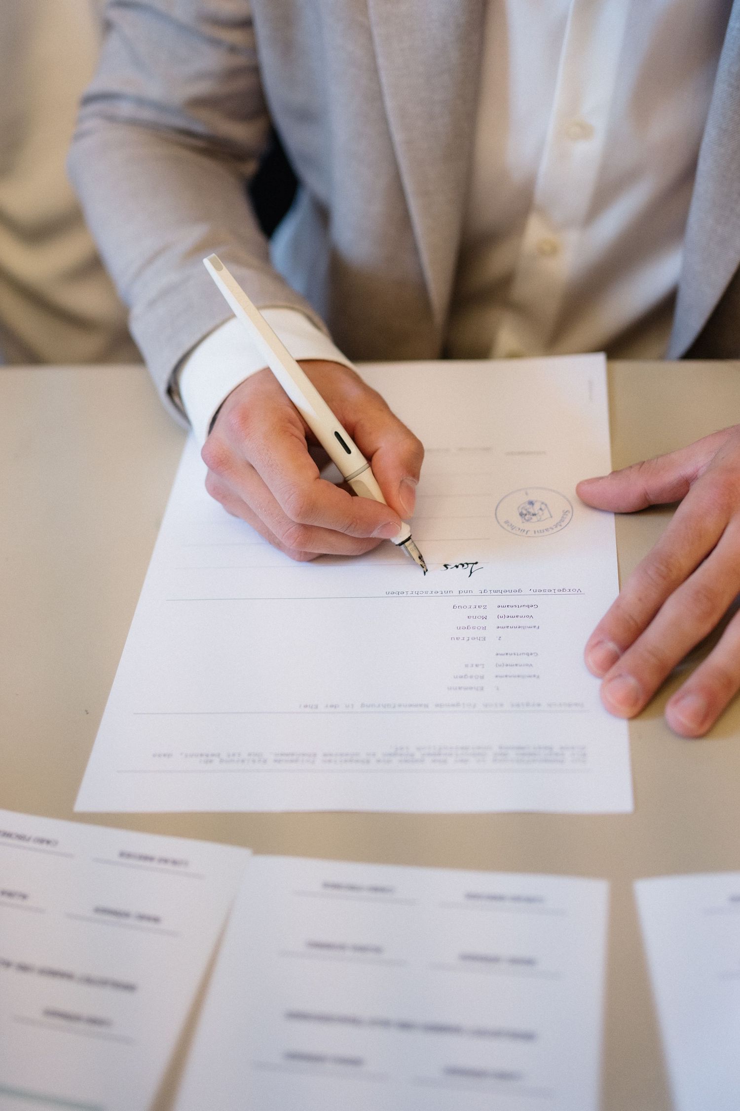 Groom in grey suit signing official wedding documents with a pen at a table.