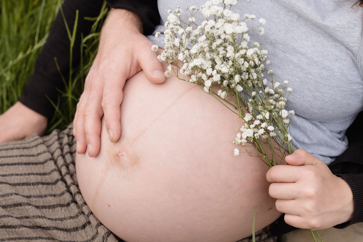 Pregnant woman holding white baby's breath flowers against her bare belly outdoors in green grass.